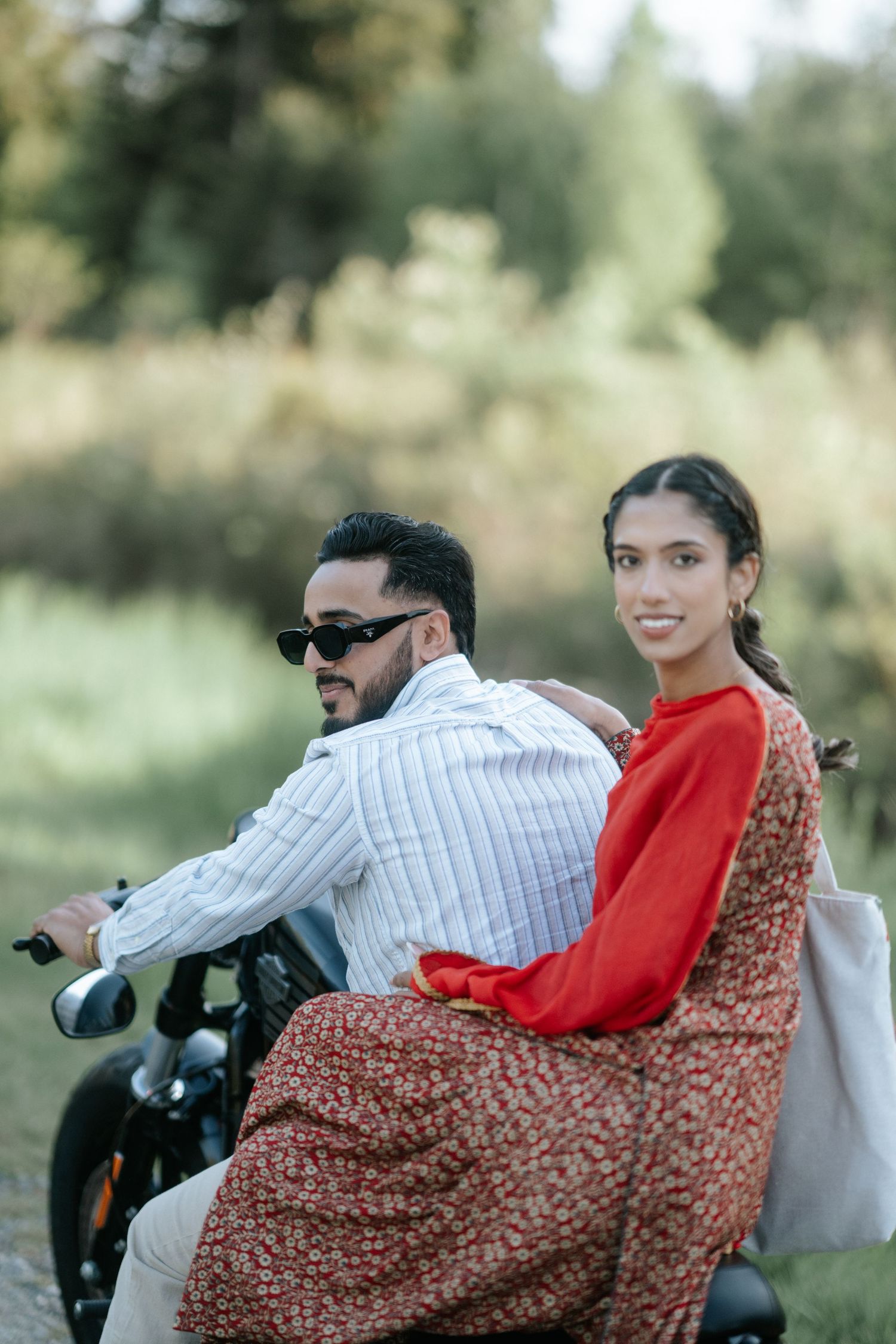 A group of people sit on a motorcycle in a casual outdoor setting with greenery in the background.