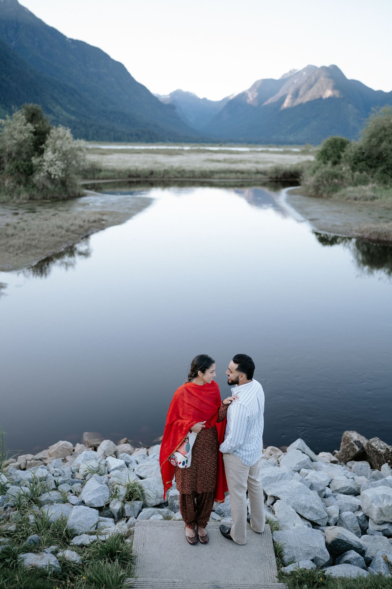 Two people sit together on rocky shoreline overlooking a serene mountain lake at dusk.