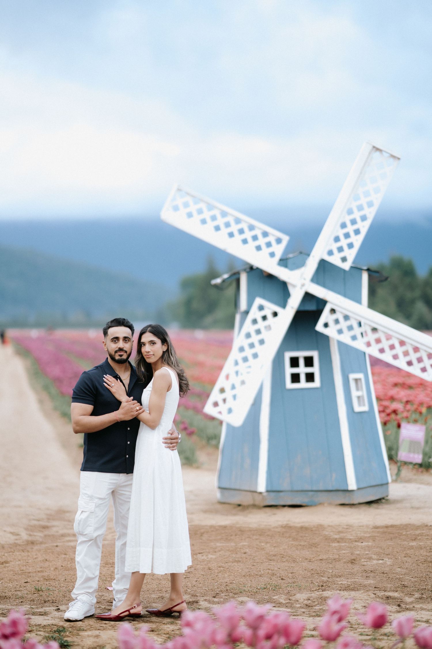 A couple poses together in front of decorative blue windmills surrounded by pink flowers in a romantic outdoor setting.