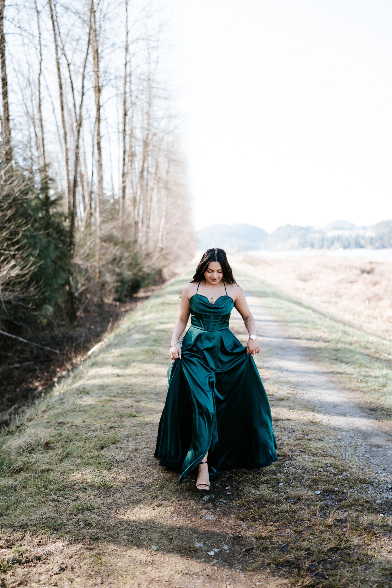 A figure in an elegant teal evening gown poses gracefully on a grassy path with trees in the background.