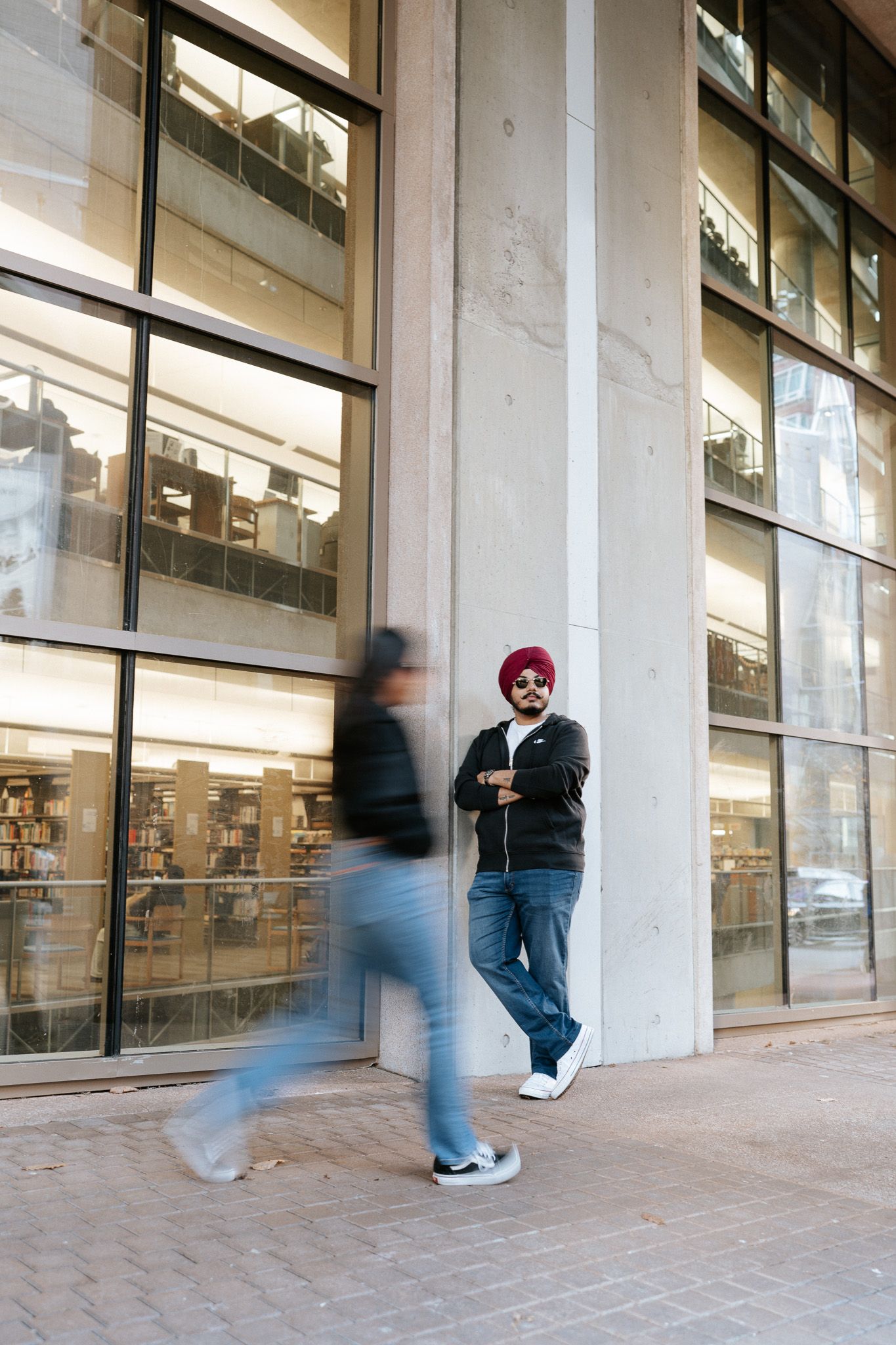 Person in a red turban and black jacket leans against modern building column while someone walks past blurred.