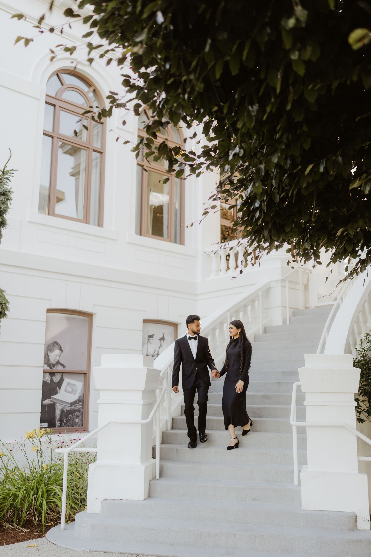 Elegant couple in formal attire walking down white stairs of a grand white building.
