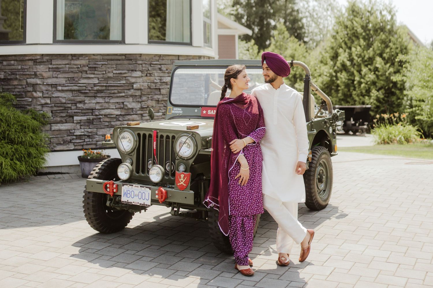 Sikh couple in traditional attire posing by a vintage green Jeep on a driveway, bride in purple and groom in white.