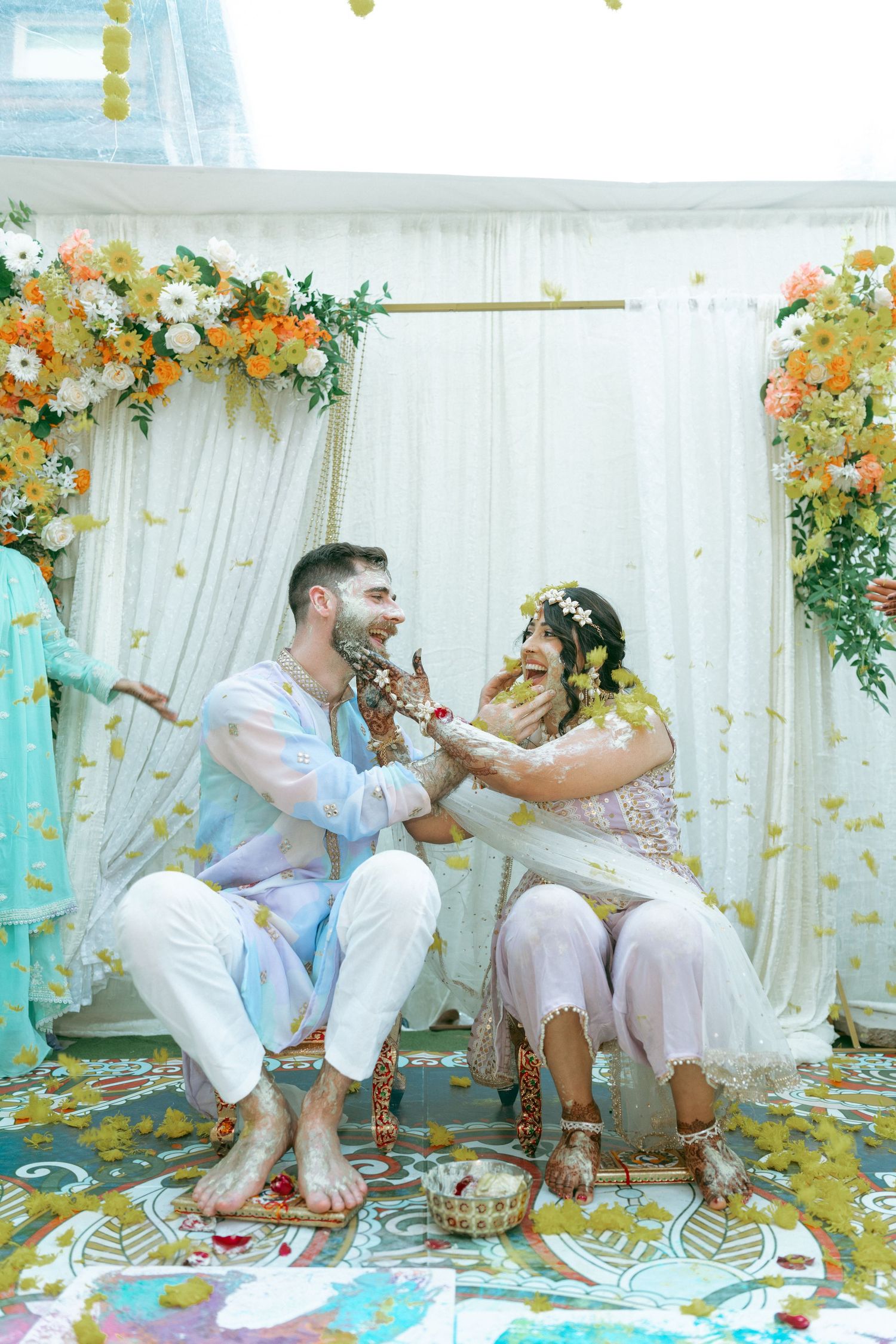 Couple joyfully applying haldi paste during Indian wedding ceremony under floral arch with marigolds.