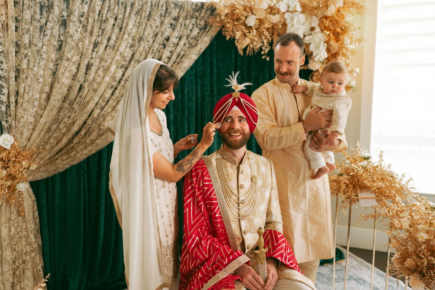 Groom in red sherwani seated during Indian wedding ceremony with family applying tilak ritual.