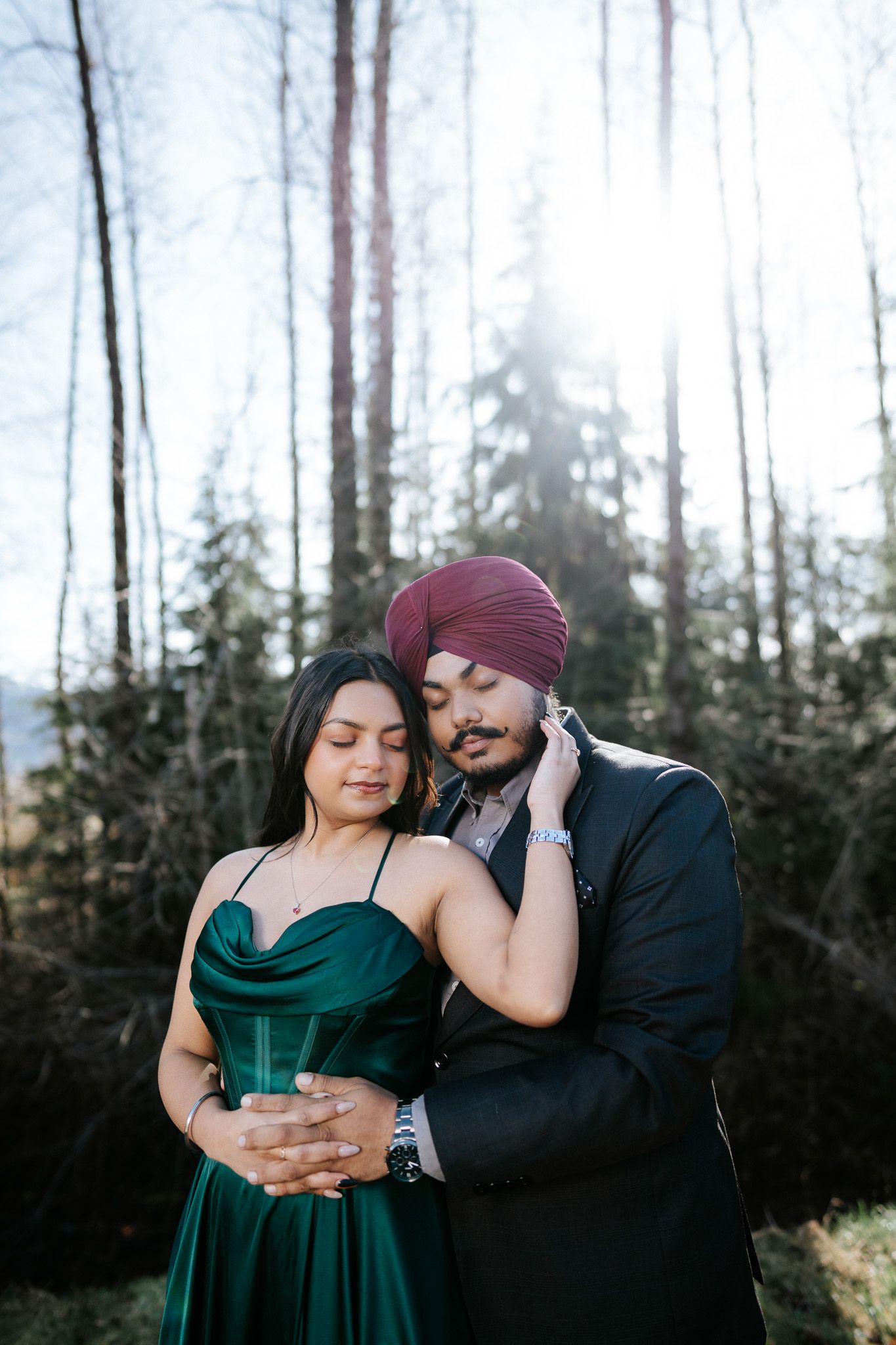 A couple shares an intimate moment together in a forest setting wearing dark clothing at sunset.