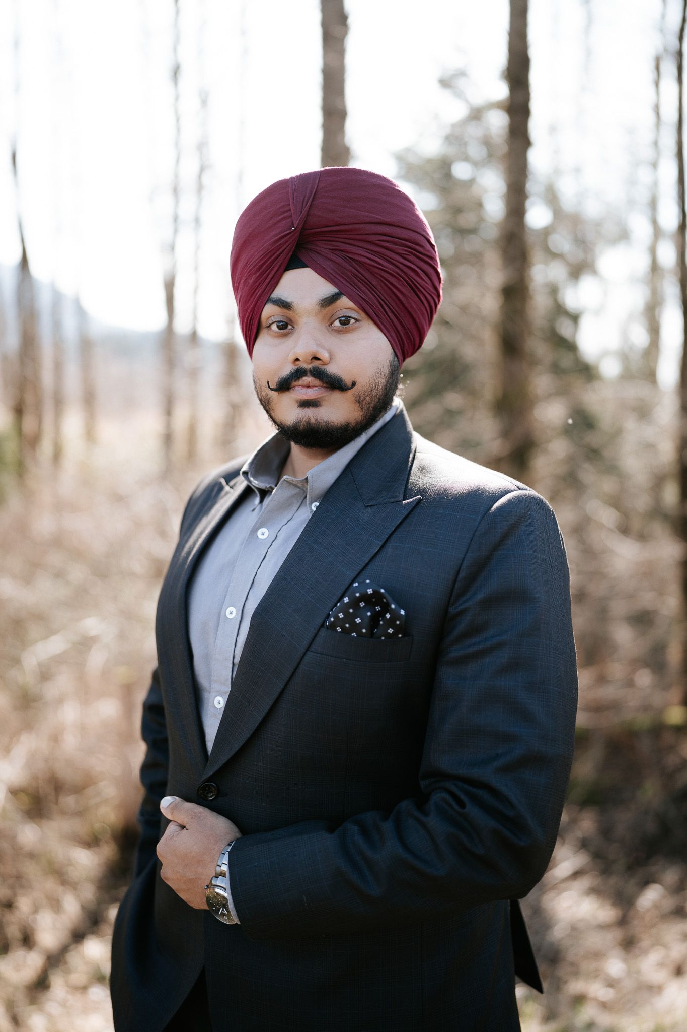 A person in a dark suit and burgundy turban poses outdoors against a natural background.