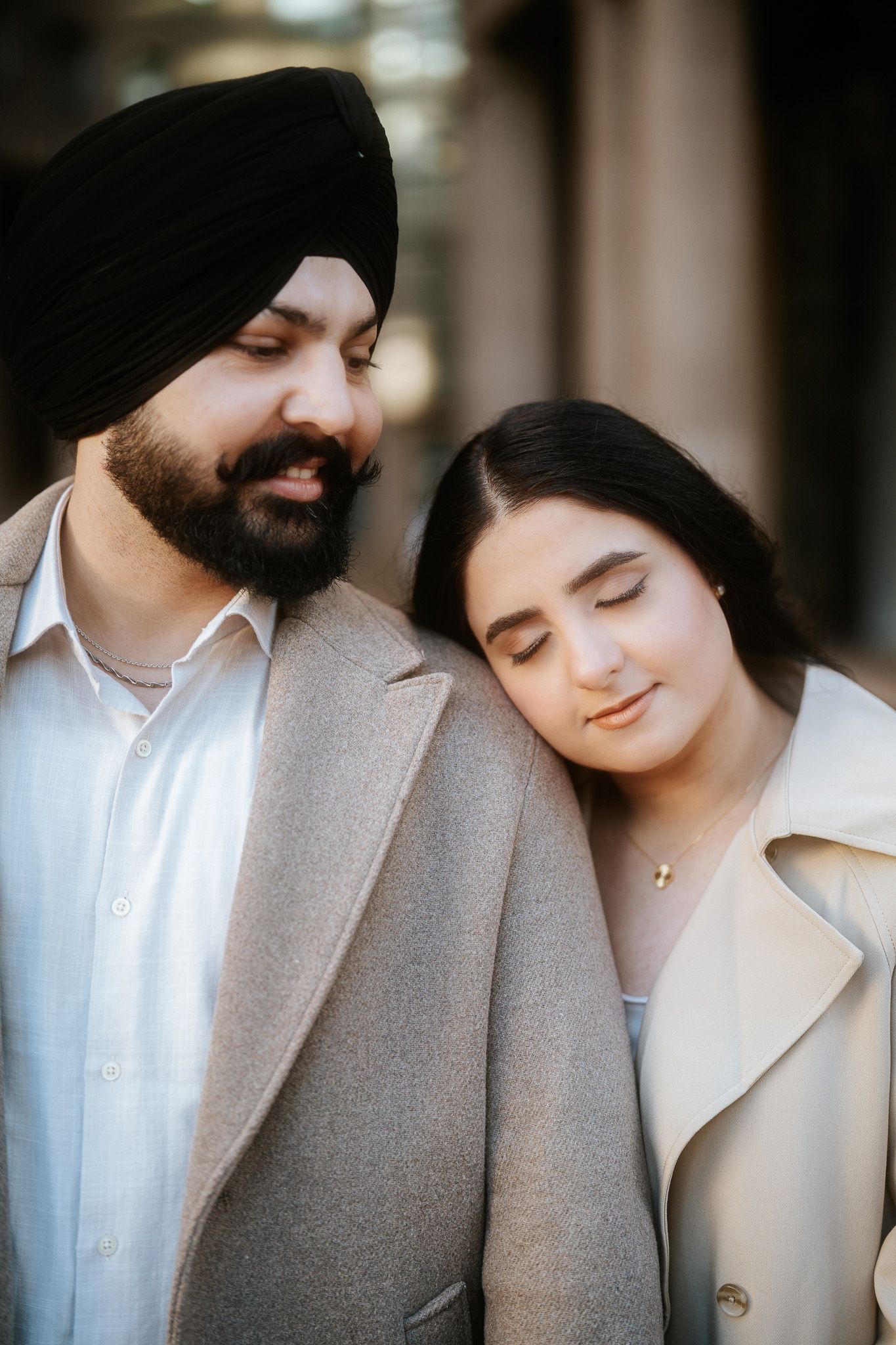 A couple in matching beige coats share an intimate moment with their eyes closed while standing close together.