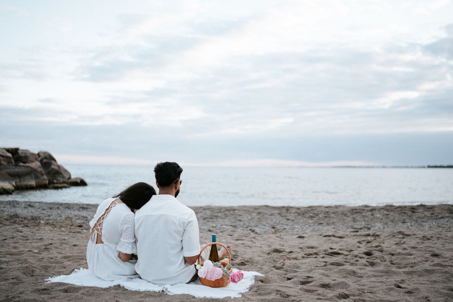 A couple sits on a white blanket having a romantic picnic by the beach at sunset with ocean and rocks in view.