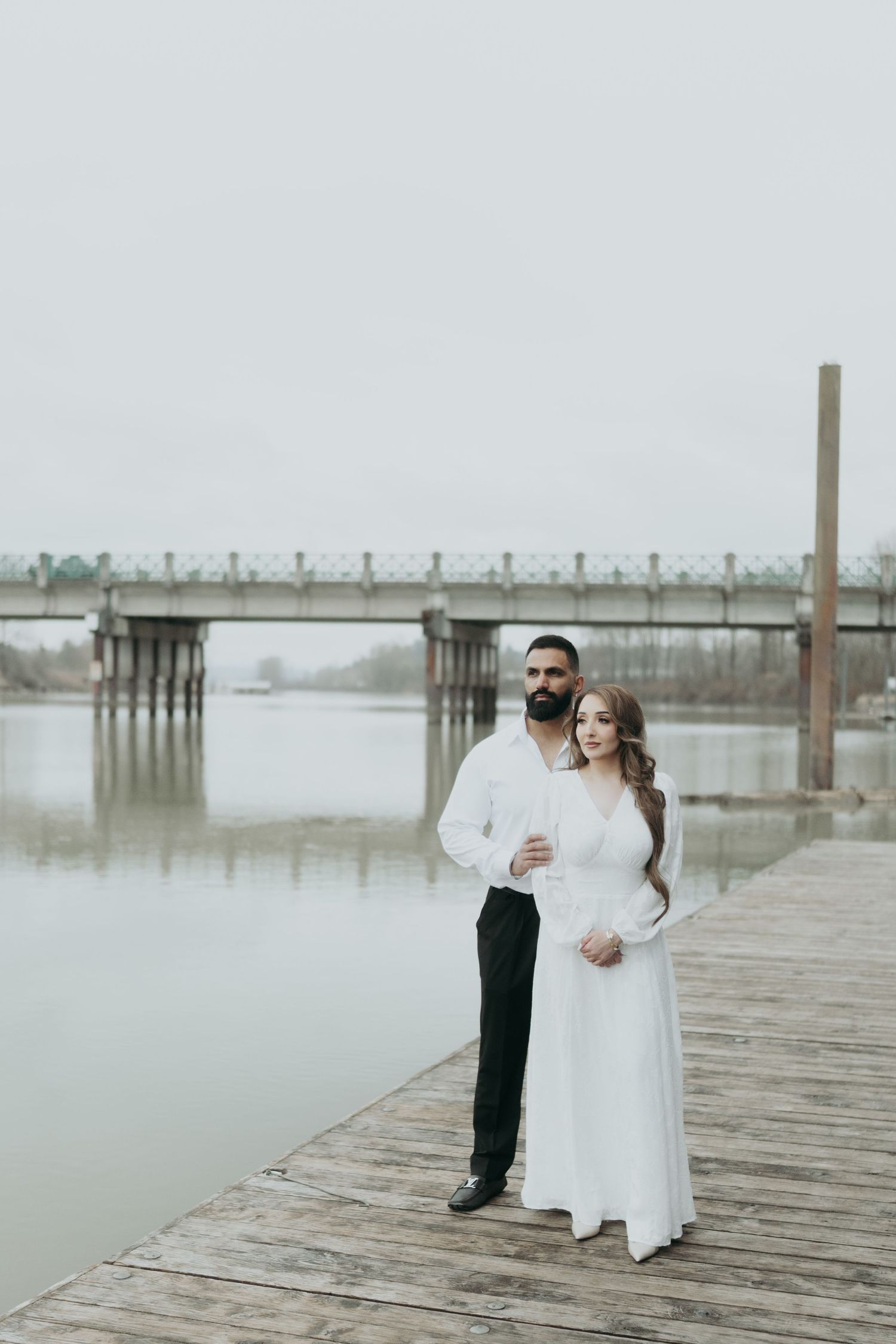 A couple poses together on a misty wooden dock overlooking a bridge and water in the distance.