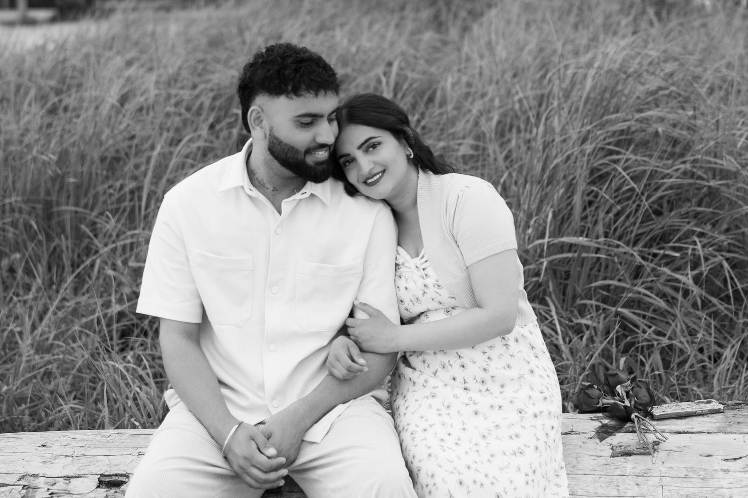 A couple in white clothing shares tender moments on a beach with tall grass in background.