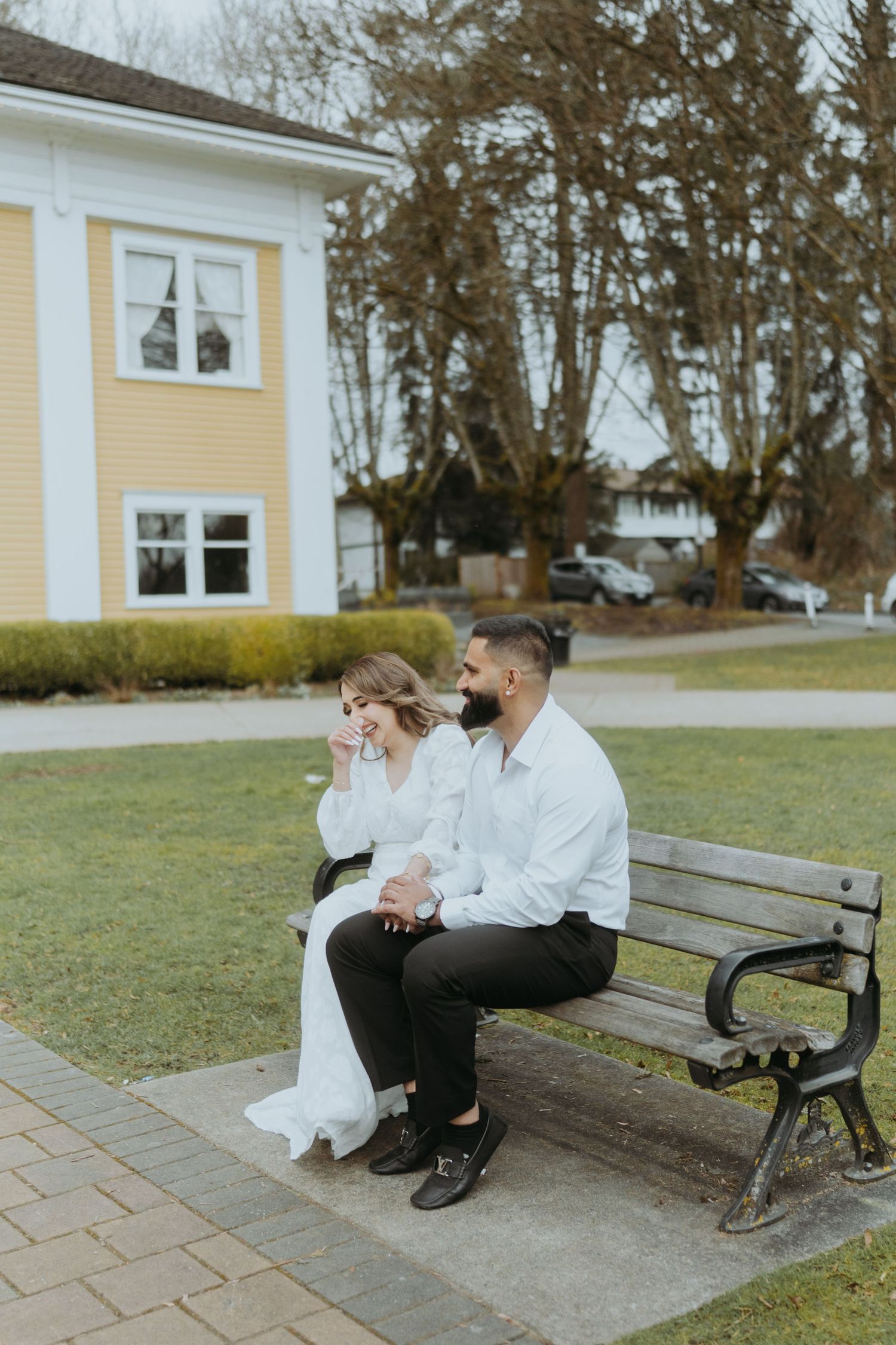 A couple sits close together on a park bench outside a yellow colonial building.