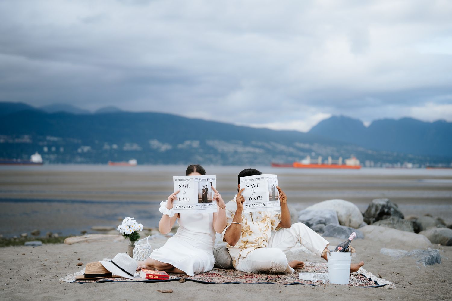 Two people sitting on a beach holding signs, with mountains and cloudy sky in the background.