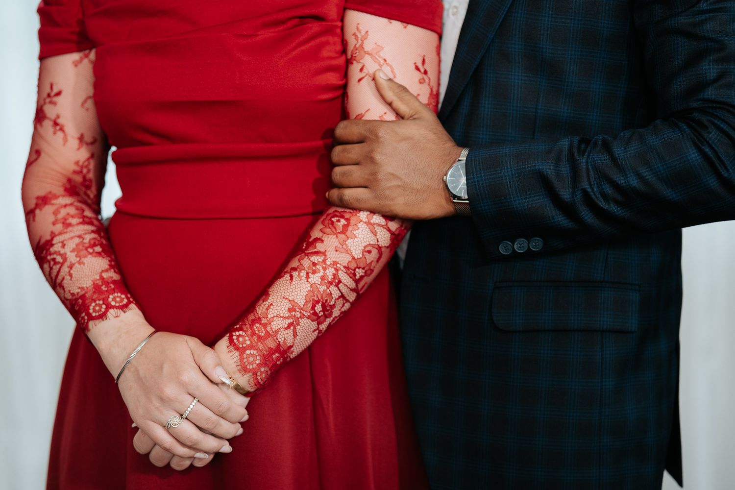 Close-up of couple holding hands, woman wearing red lace long-sleeve gown and man in navy plaid suit with silver watch.