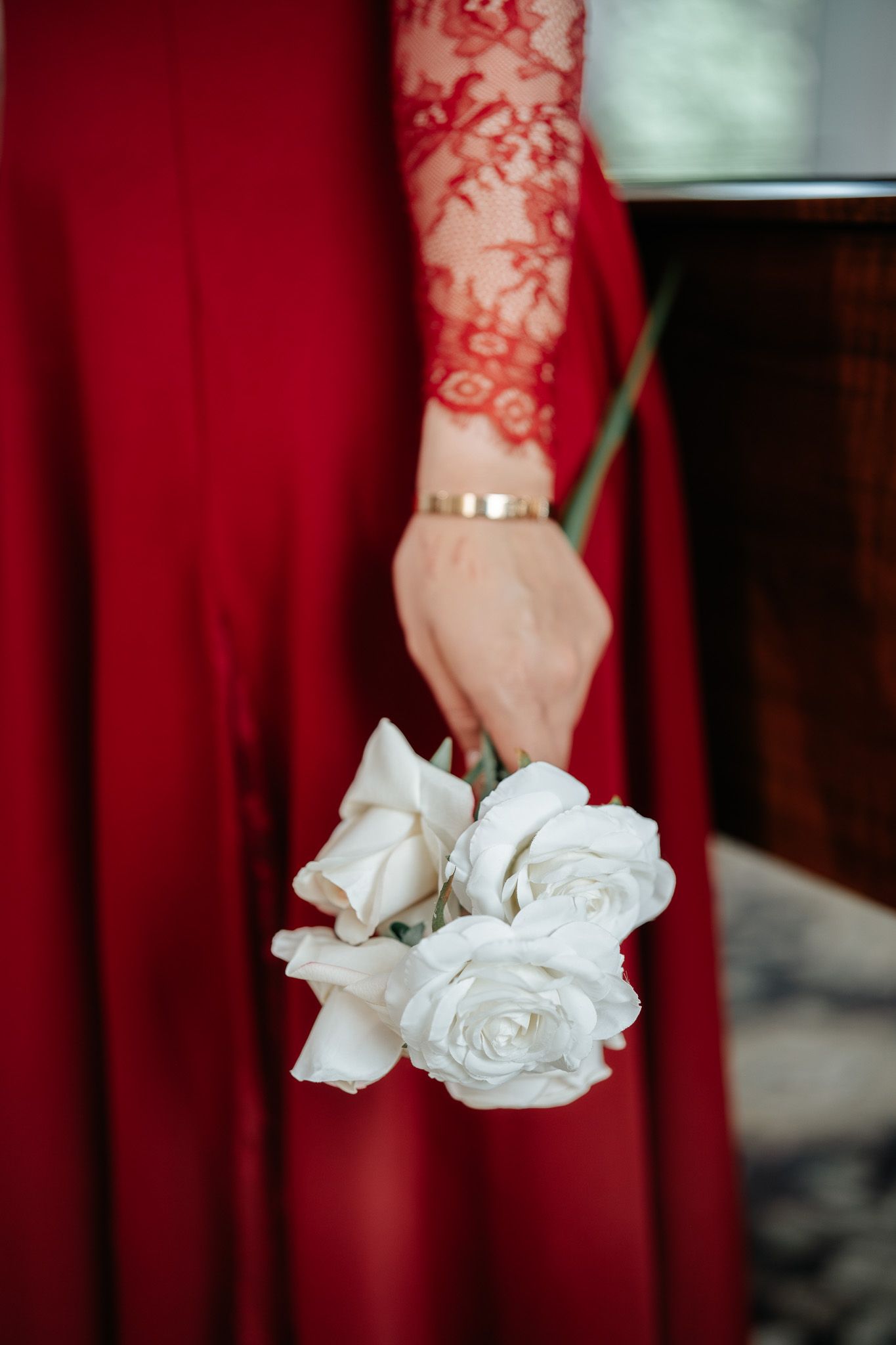 Close-up of woman in red lace gown holding small white rose bouquet with gold bracelet accent.