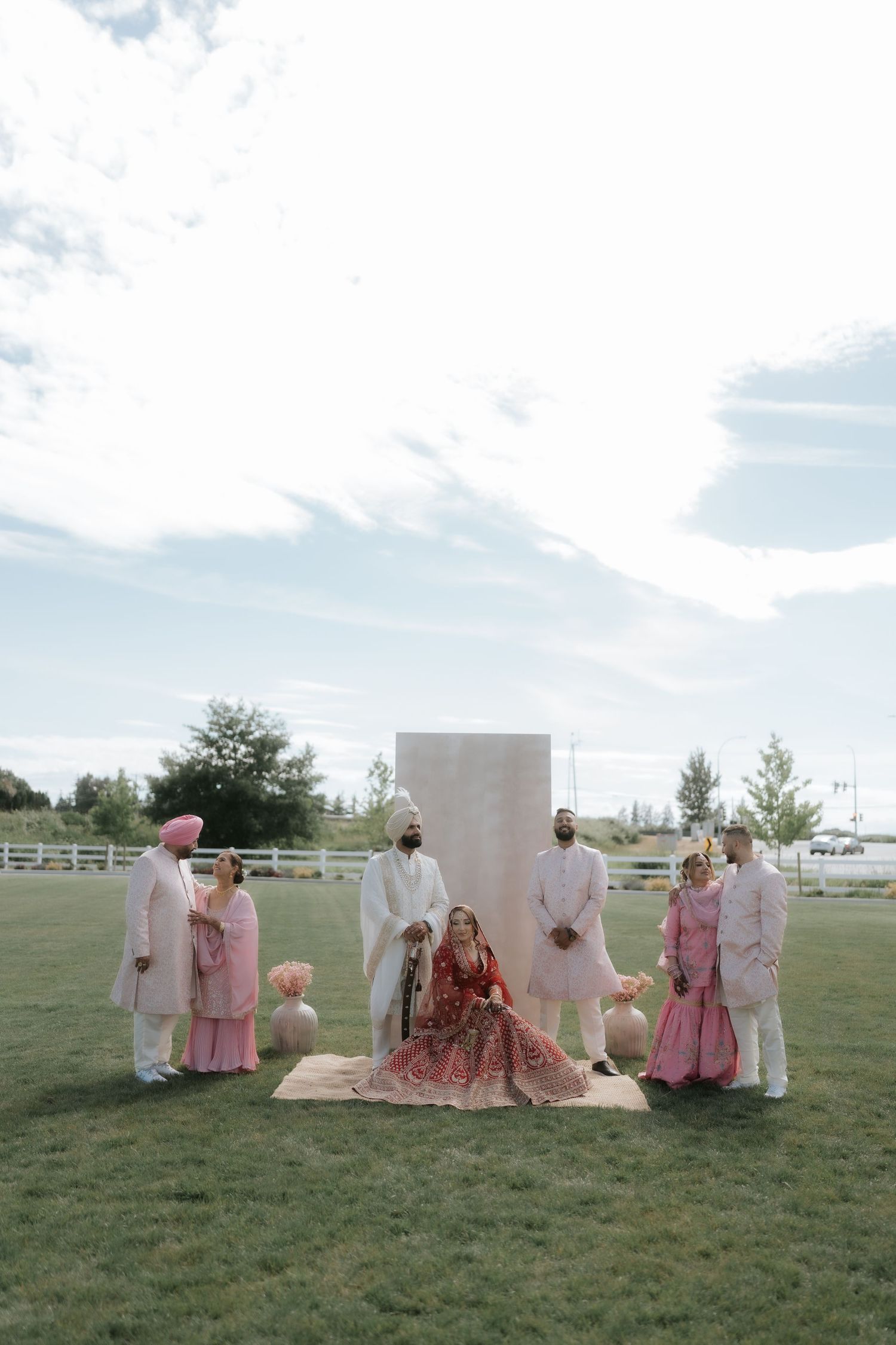 South Asian wedding party poses outdoors in traditional attire, bride in red lehenga seated center before white backdrop.