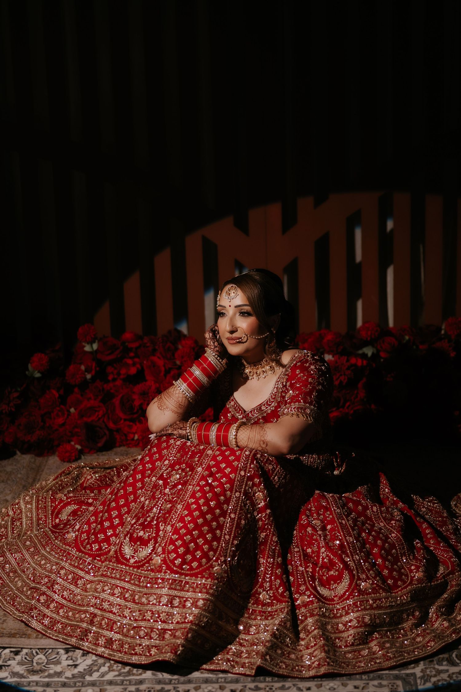 Indian bride in red embroidered lehenga with gold jewelry poses elegantly surrounded by red roses.