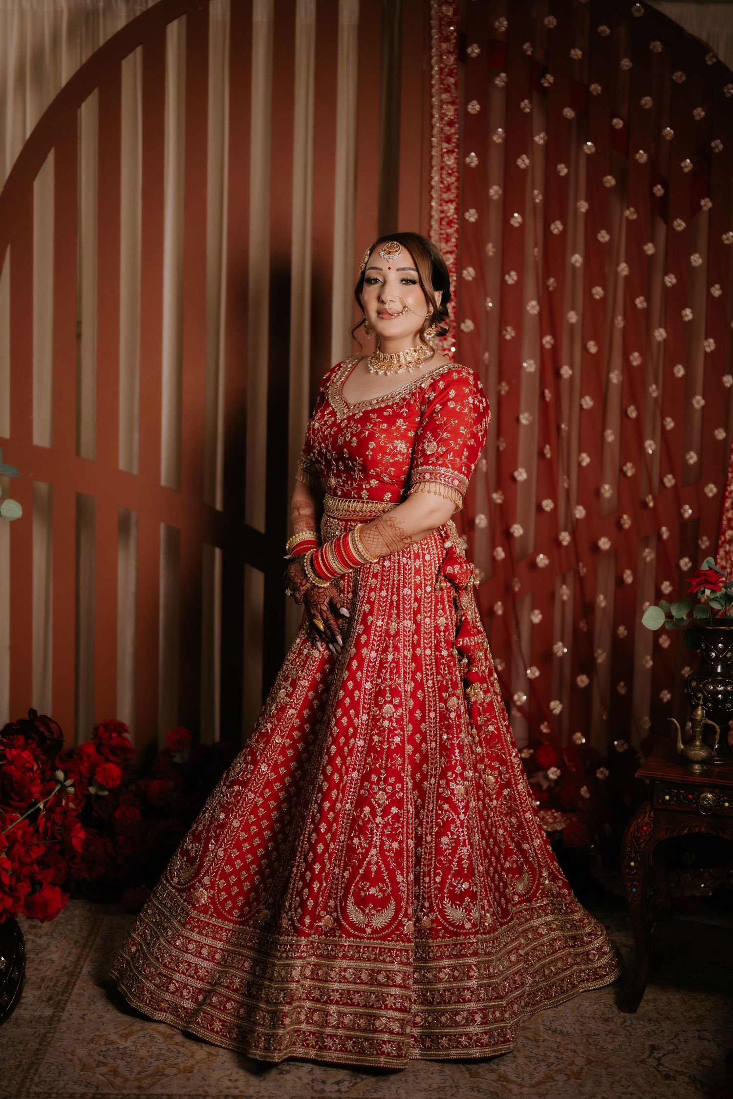 A bride in a red embroidered lehenga with gold jewelry poses elegantly against a decorative red backdrop.
