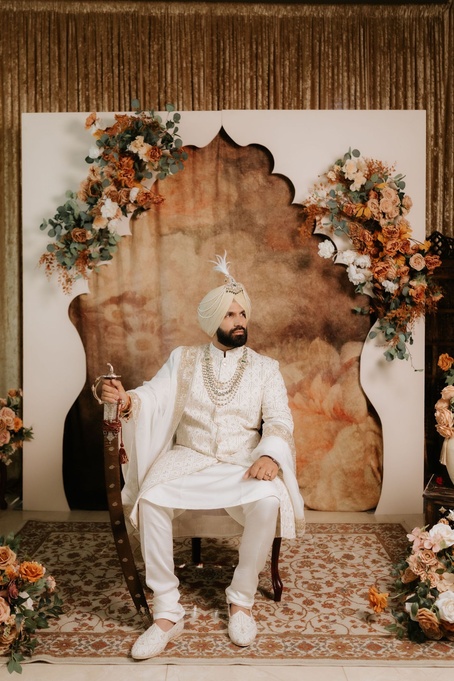 Sikh groom in white sherwani and turban seated on ornate chair with floral backdrop at wedding ceremony.