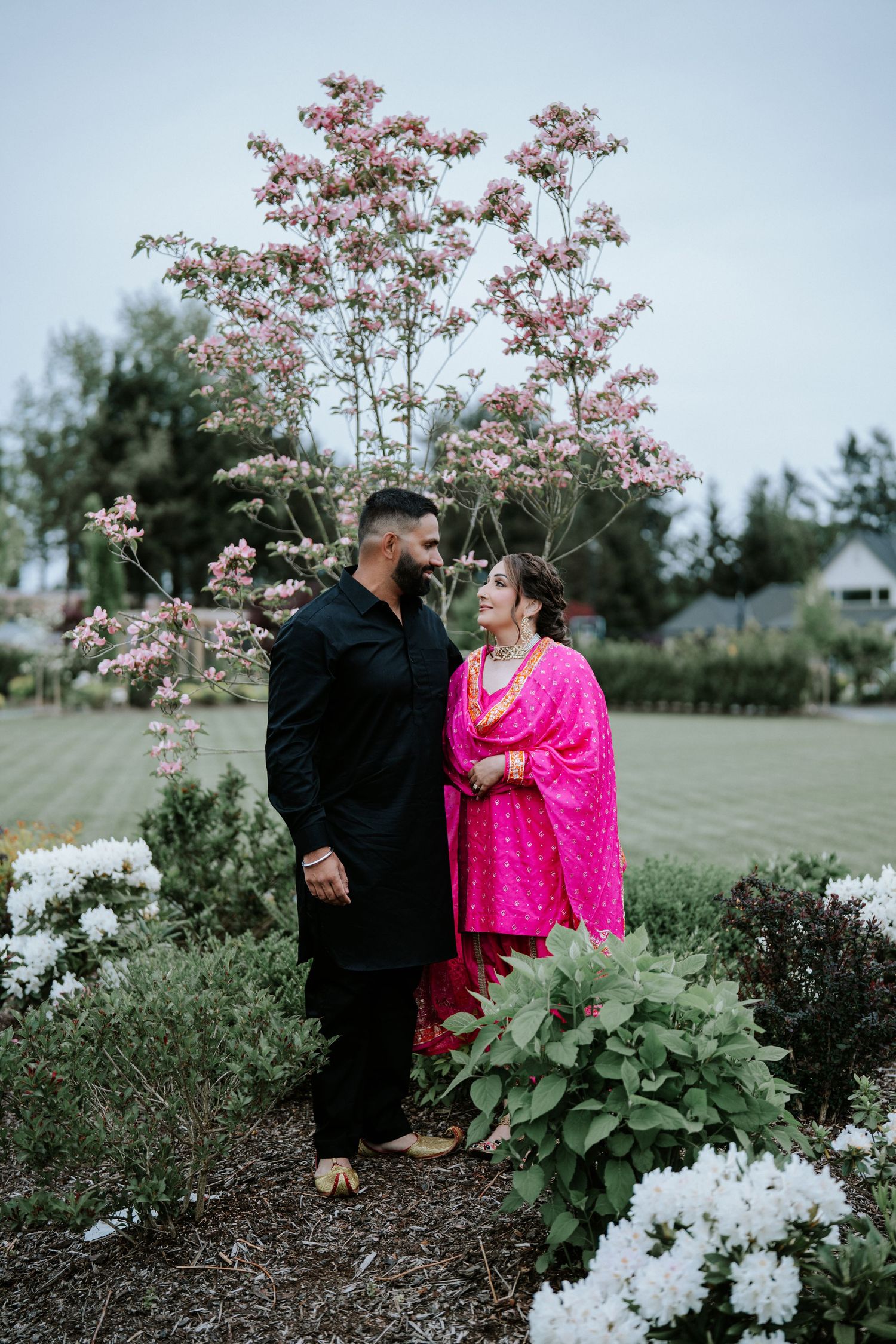 Couple in traditional South Asian attire standing in a garden with blooming pink flowers and white hydrangeas.