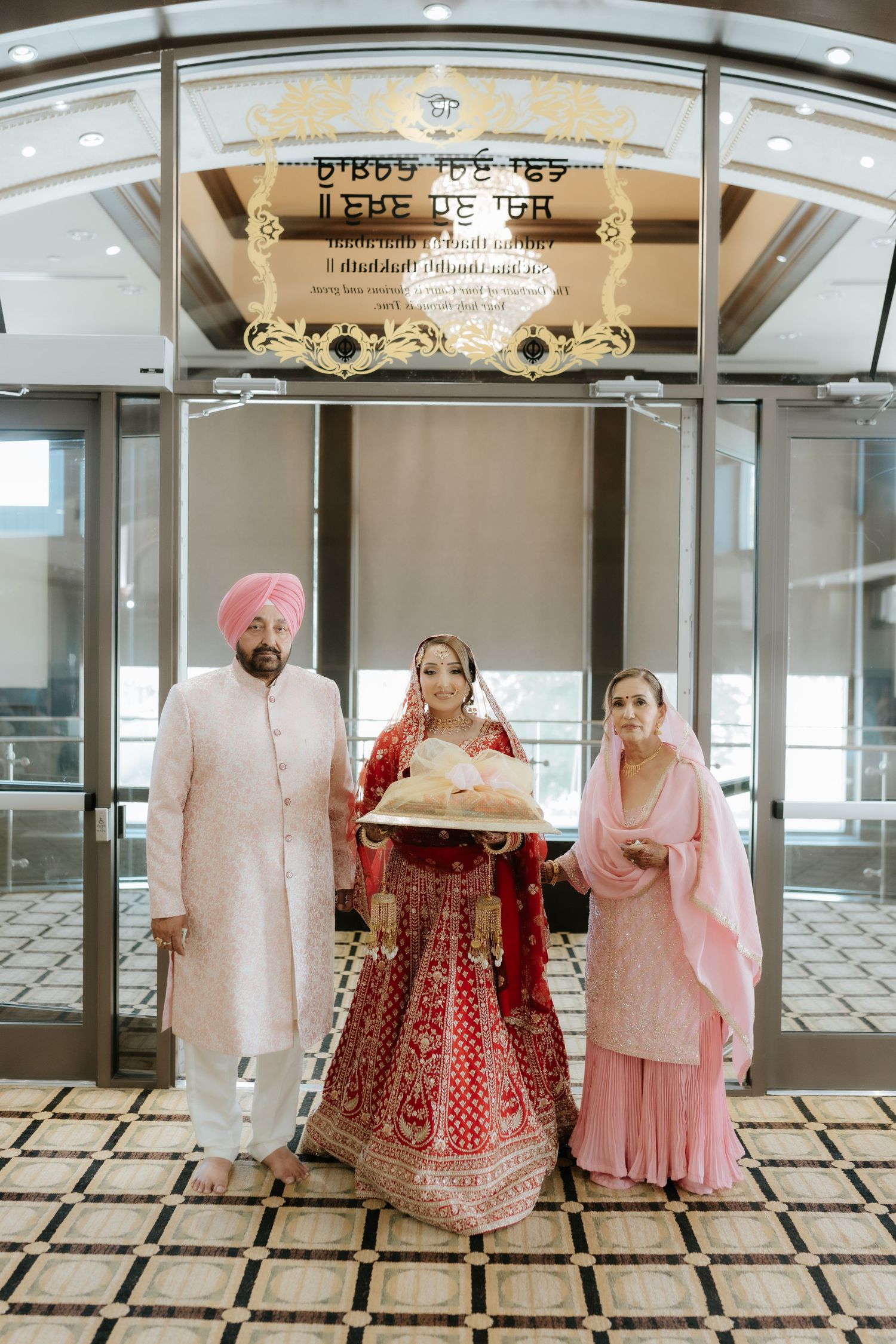 Sikh bride in red lehenga stands with family outside gurdwara entrance for traditional wedding portrait.