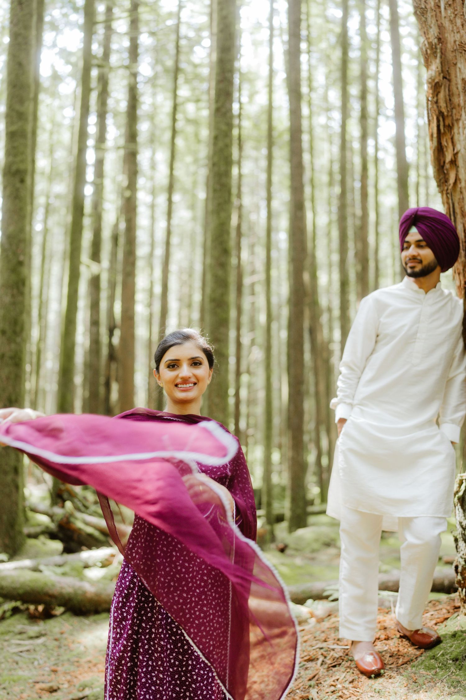 Sikh couple in traditional attire posing in a forest, woman twirling purple dupatta while man in white kurta watches.