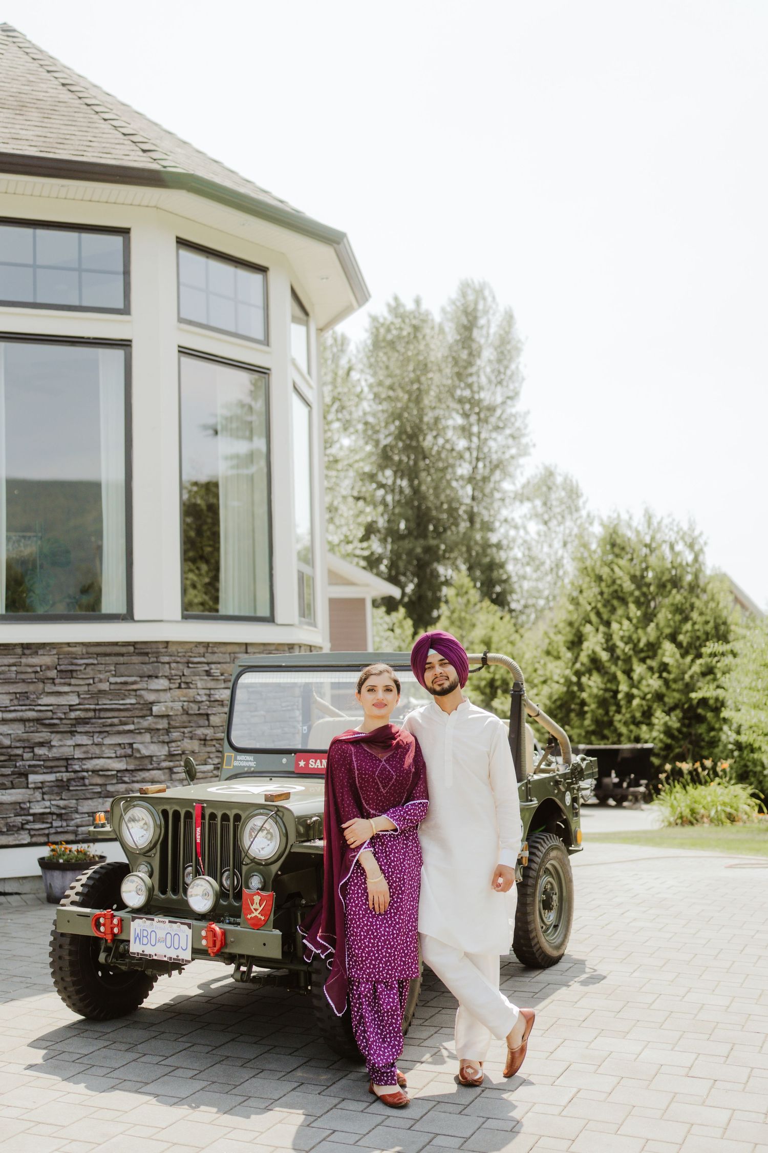 Sikh couple posing by vintage military jeep outside stone building, wearing coordinated purple and white outfits.