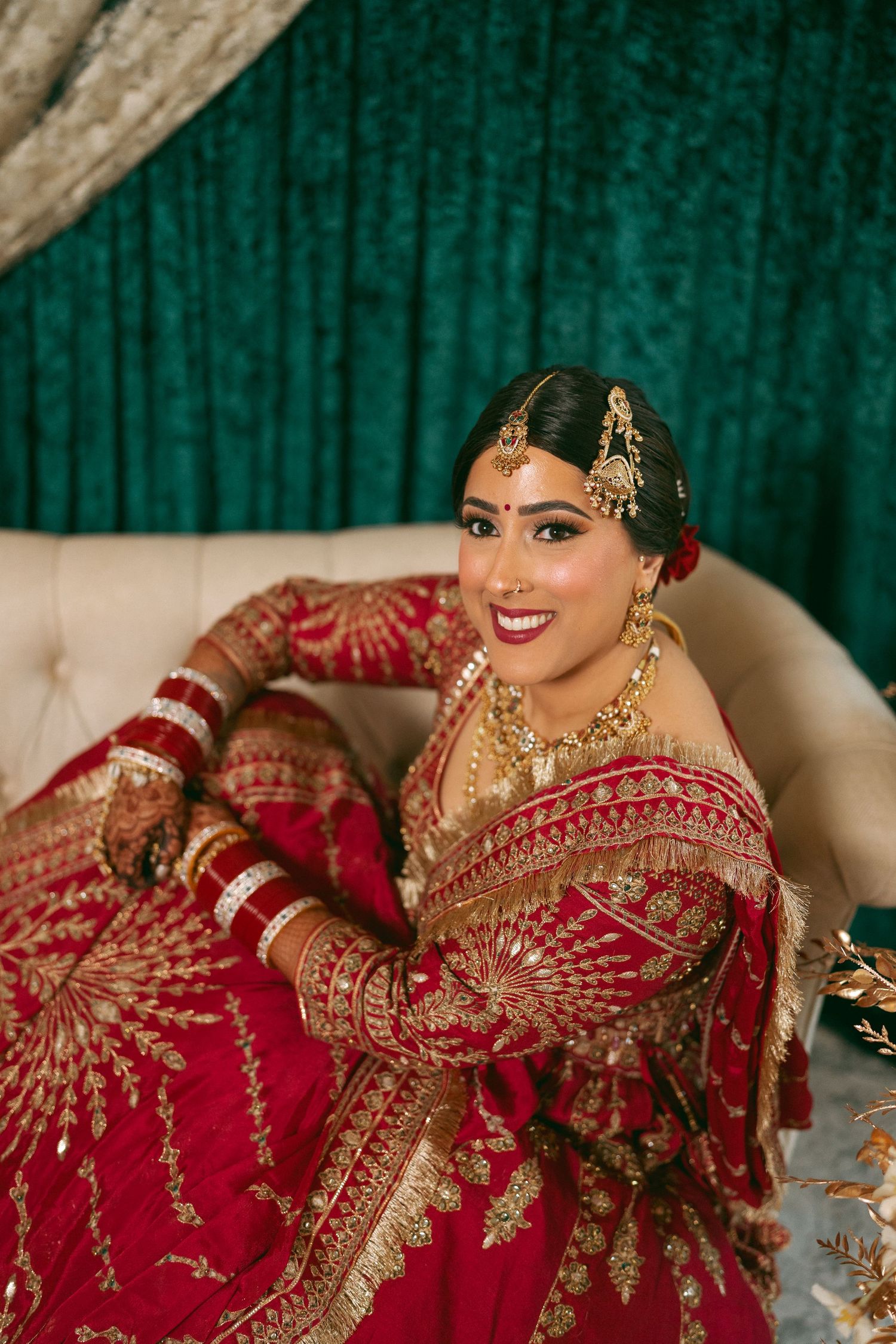 Indian bride in red and gold lehenga with traditional jewelry smiles gracefully on a cream sofa.