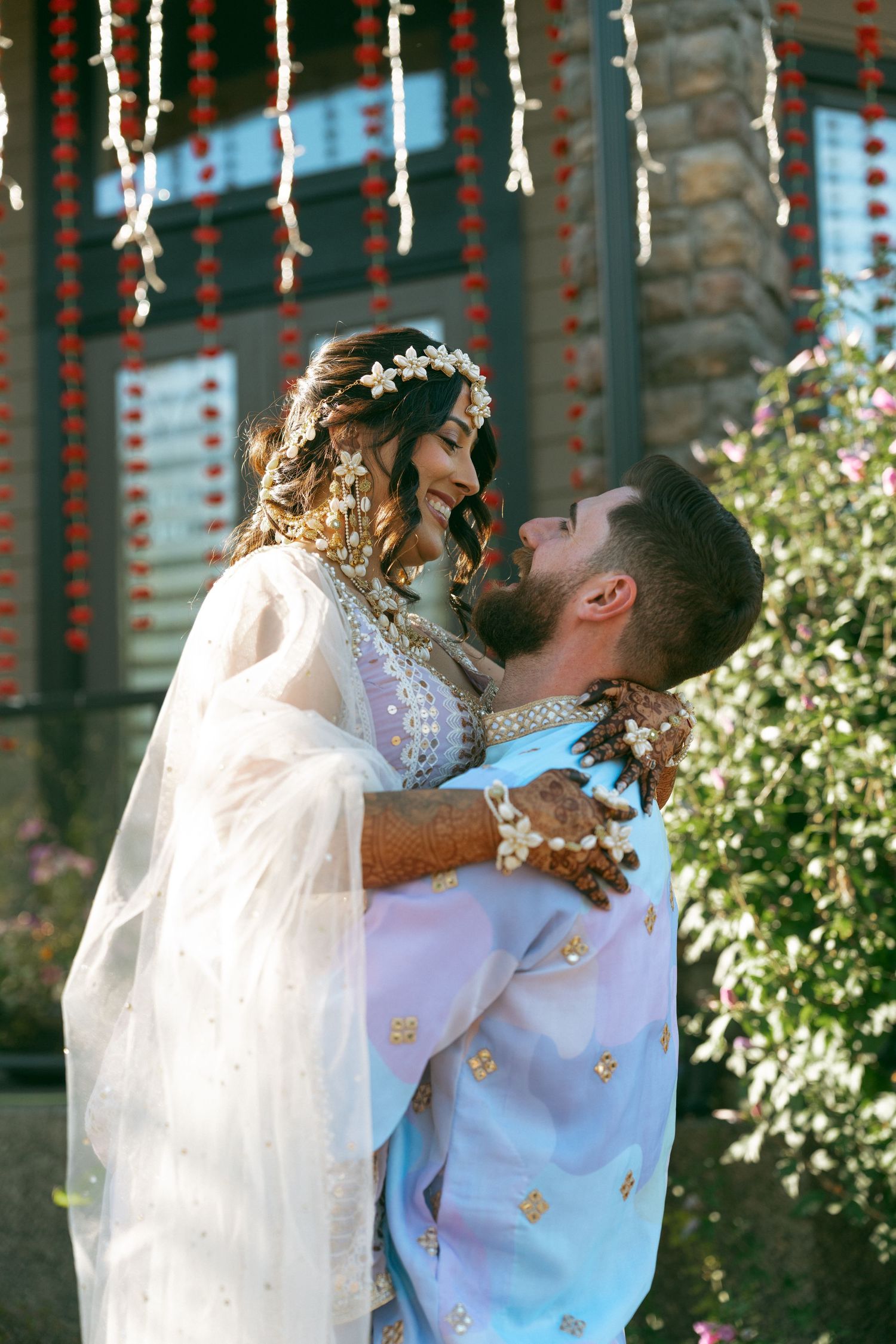 A couple in traditional South Asian wedding attire embrace outdoors, adorned with floral decorations and festive hanging garlands.