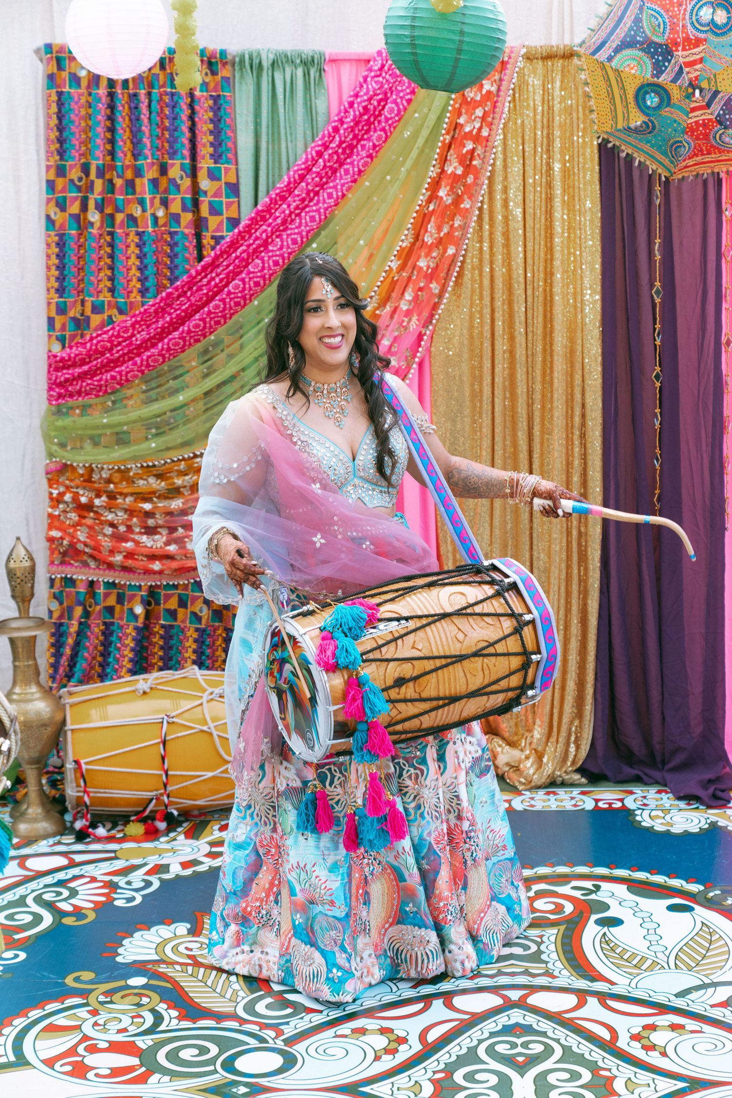 Woman in colorful lehenga playing dhol drum at vibrant Indian celebration with decorative backdrops.