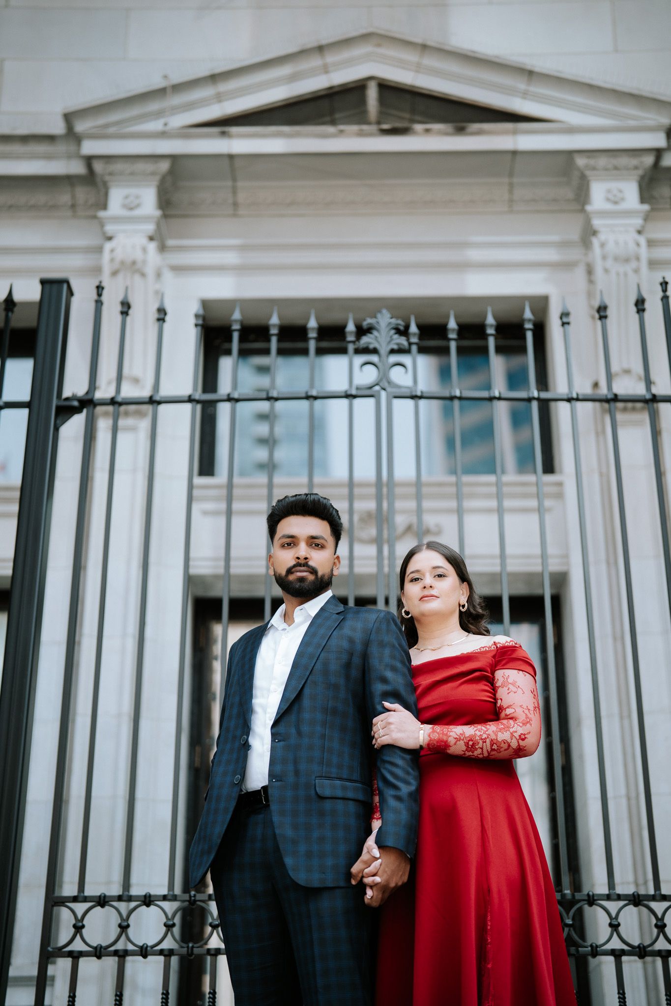 Elegant couple posing before ornate iron gates, man in plaid suit, woman in red off-shoulder gown.