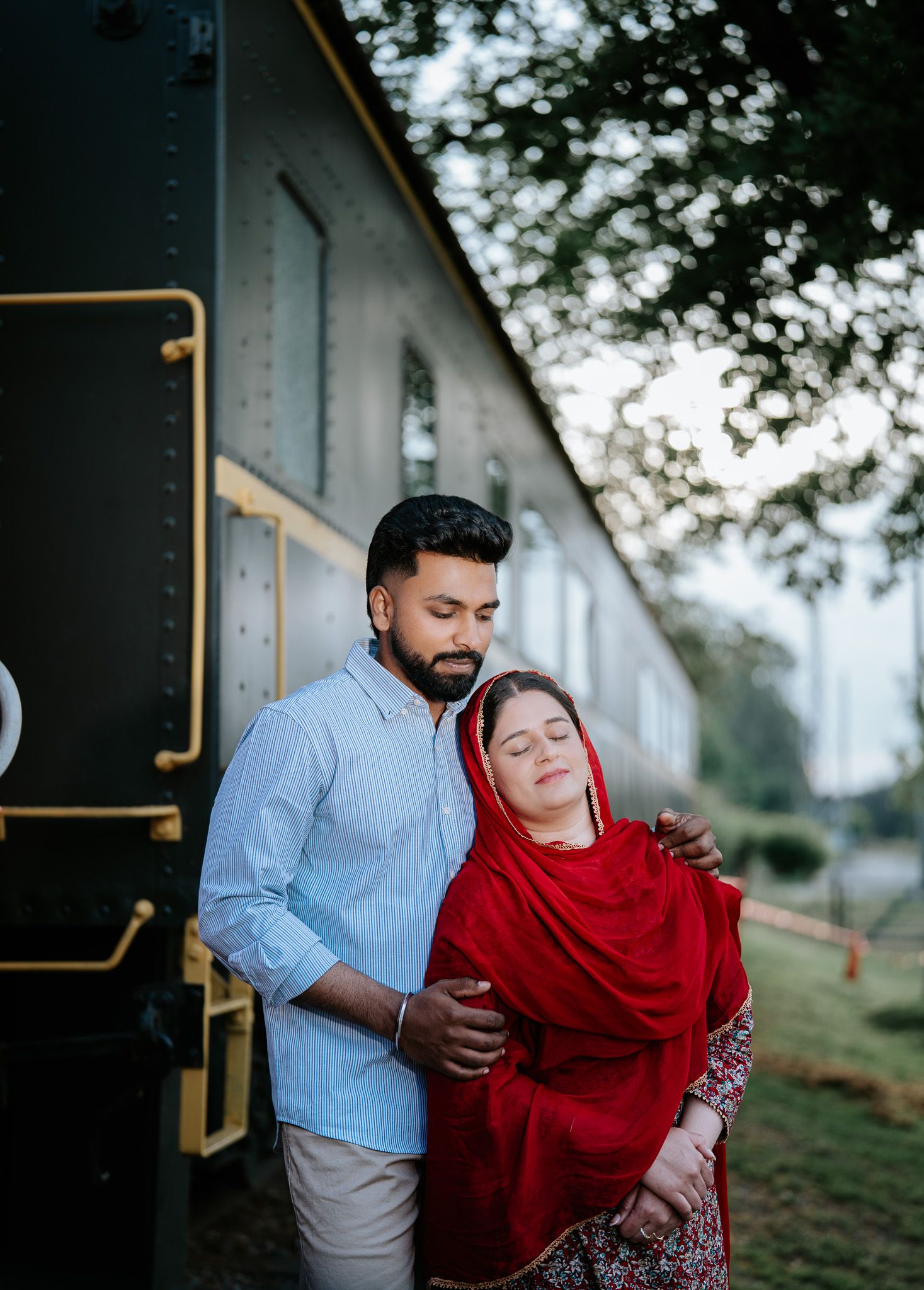 Couple embracing near a vintage train, woman in red dupatta resting on man's shoulder in a romantic outdoor portrait.