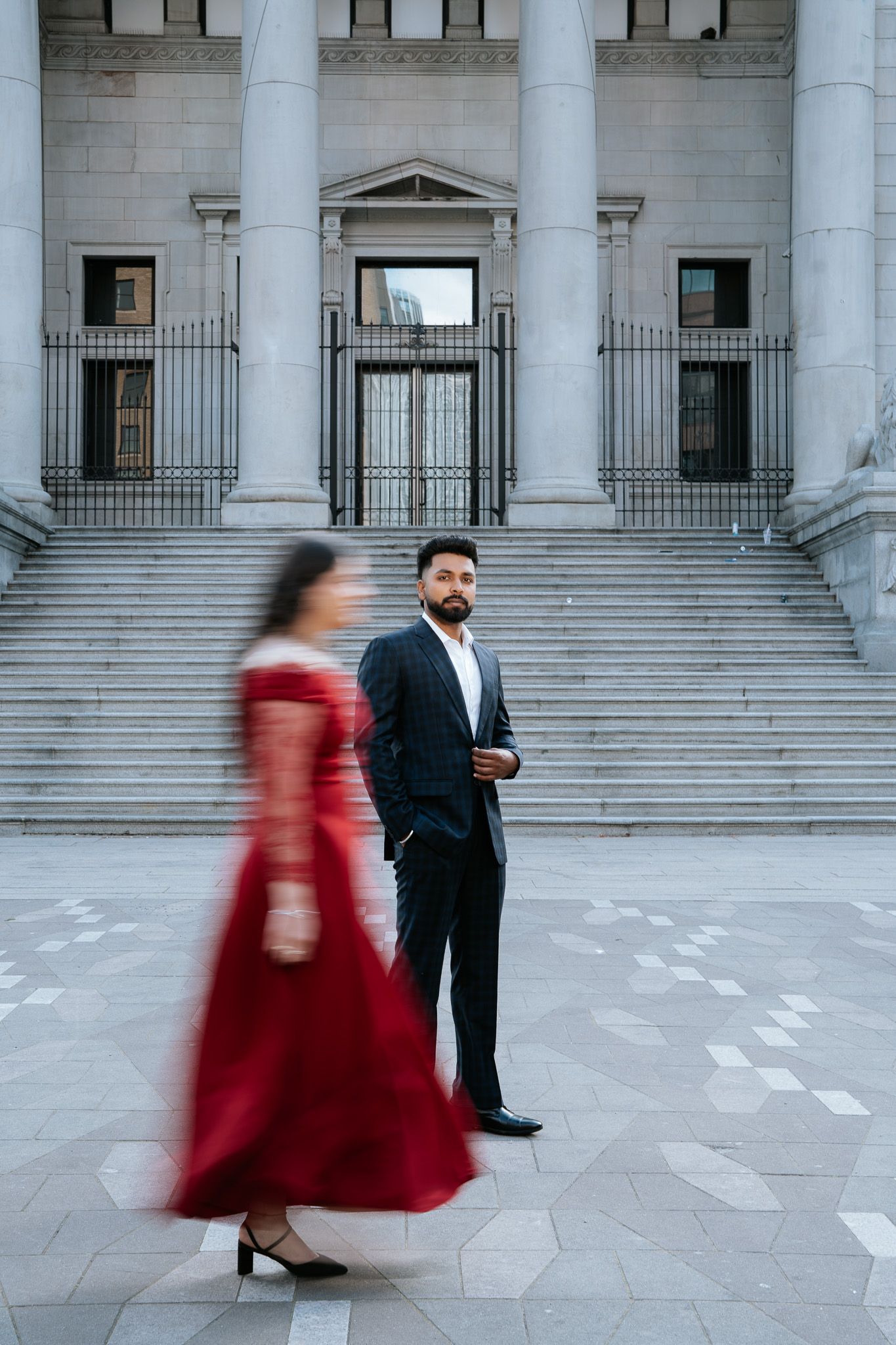 A man in a black suit stands before grand stone steps as a woman in a red dress walks past in motion blur.
