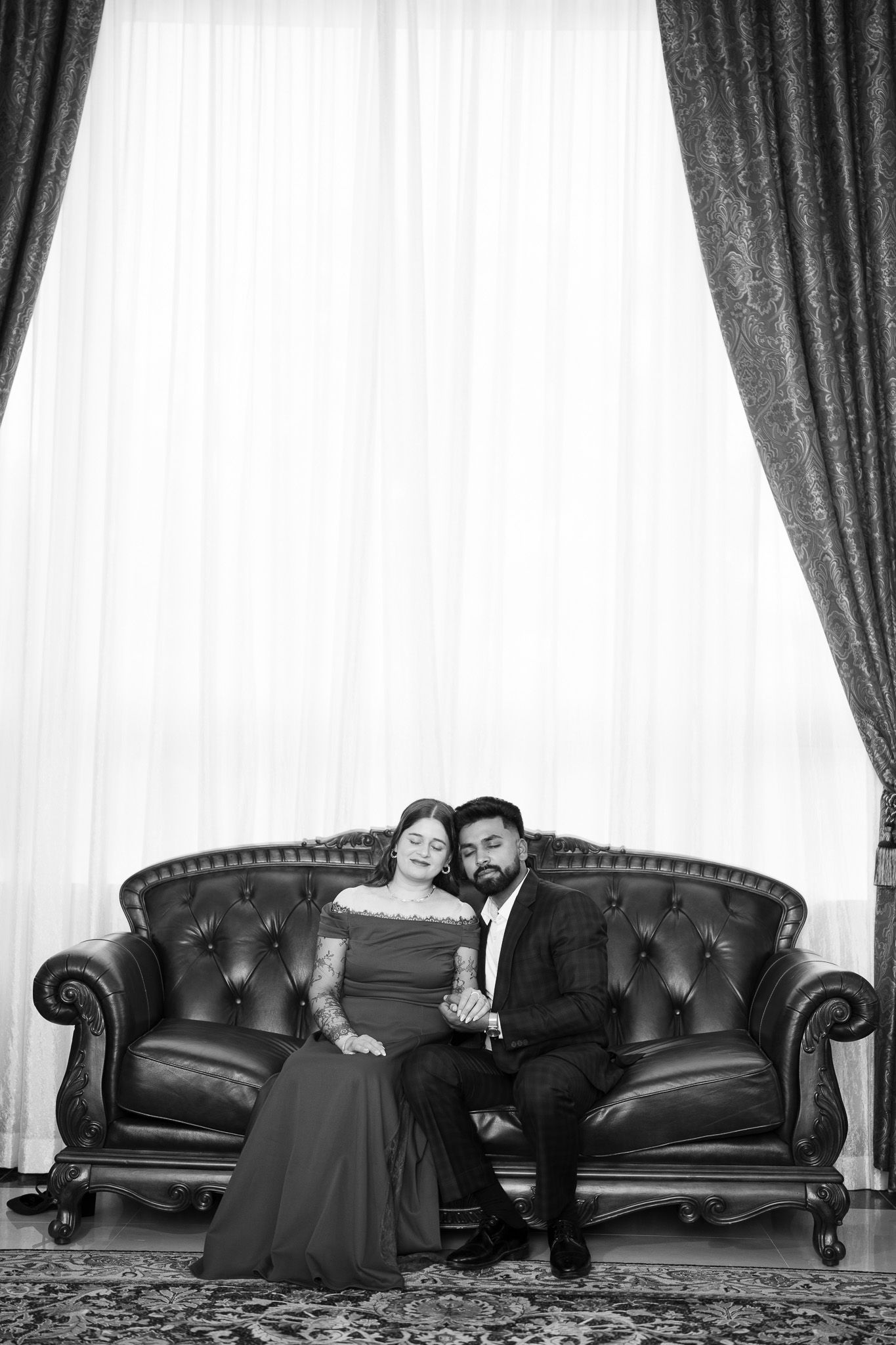 Elegant couple in formal attire sitting on tufted leather sofa in black and white portrait with draped curtains.