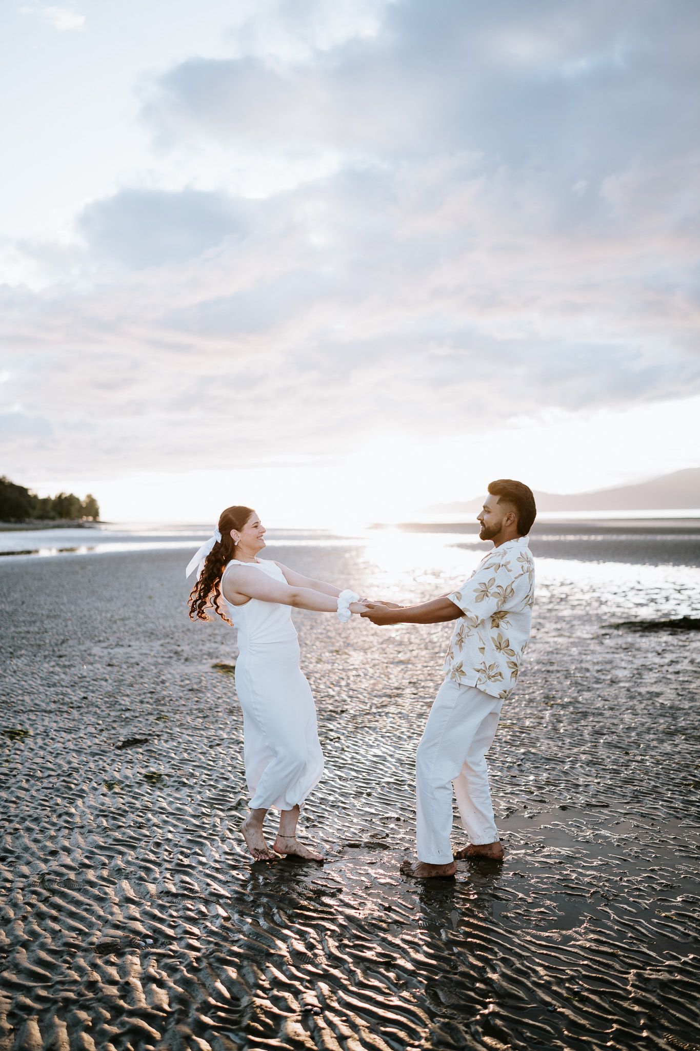 Couple in white holding hands on a reflective beach at sunset with ocean and cloudy sky backdrop.