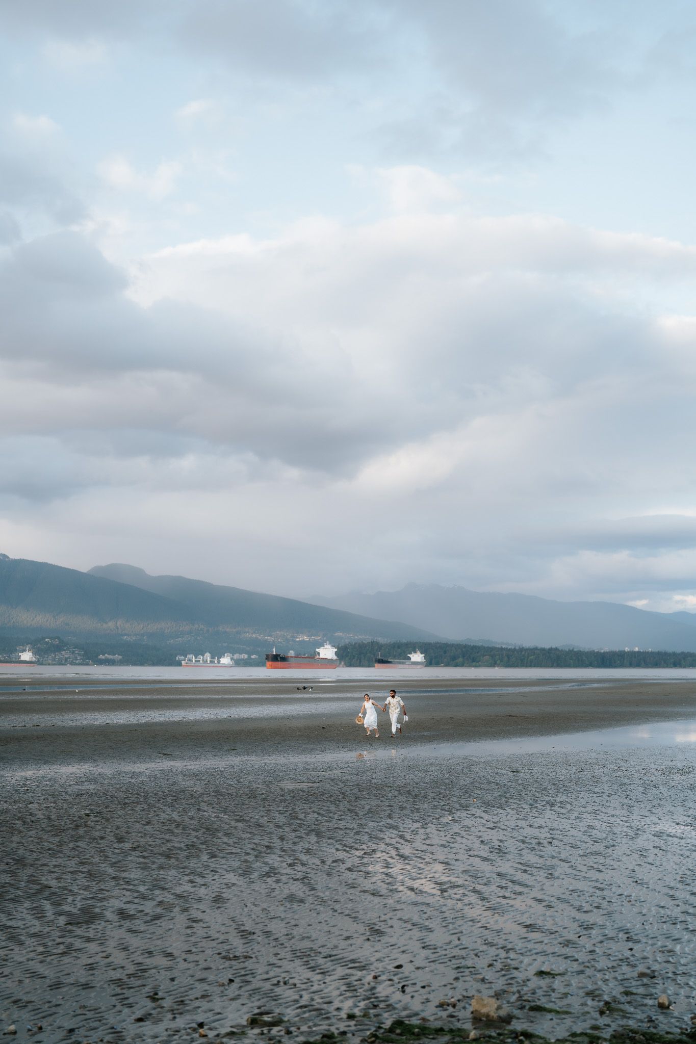 Couple in white walking on vast tidal flats with mountains and ships visible across the water.