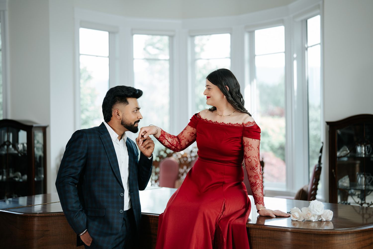 A smiling couple in formal attire share a romantic moment by a grand piano, man kissing womans hand.