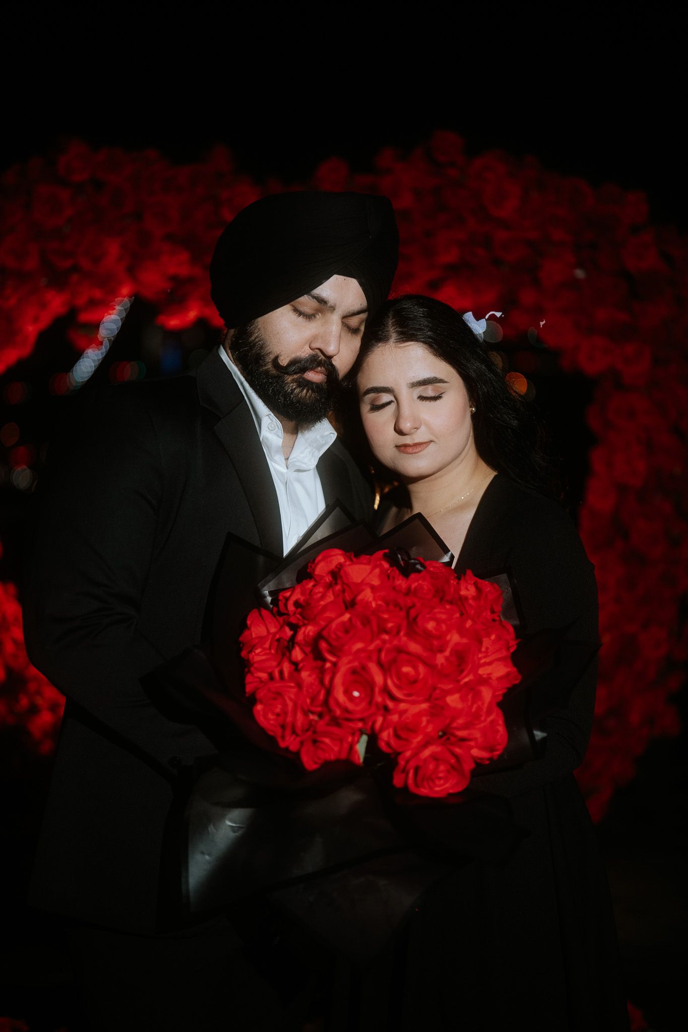 A romantic couple poses against a backdrop of red roses while holding a vibrant bouquet of red flowers.