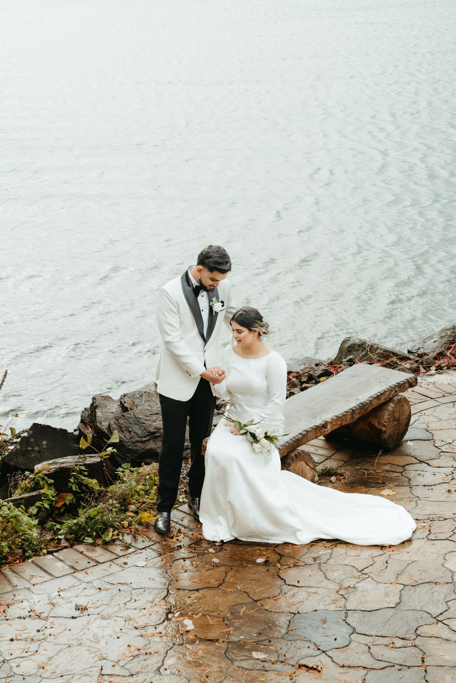 A couple in wedding attire sits together on stone steps near a lake on their special day.