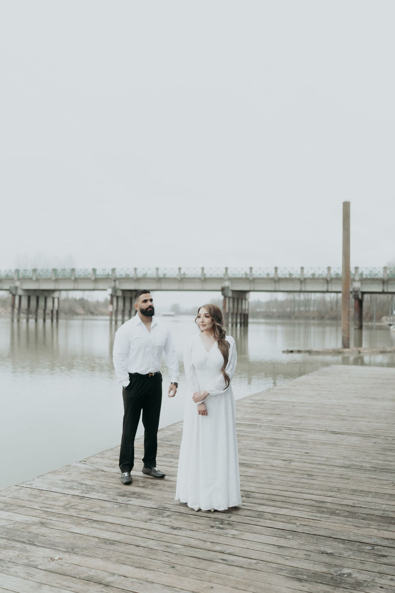 A couple in contrasting black and white outfits poses on a misty waterfront dock.