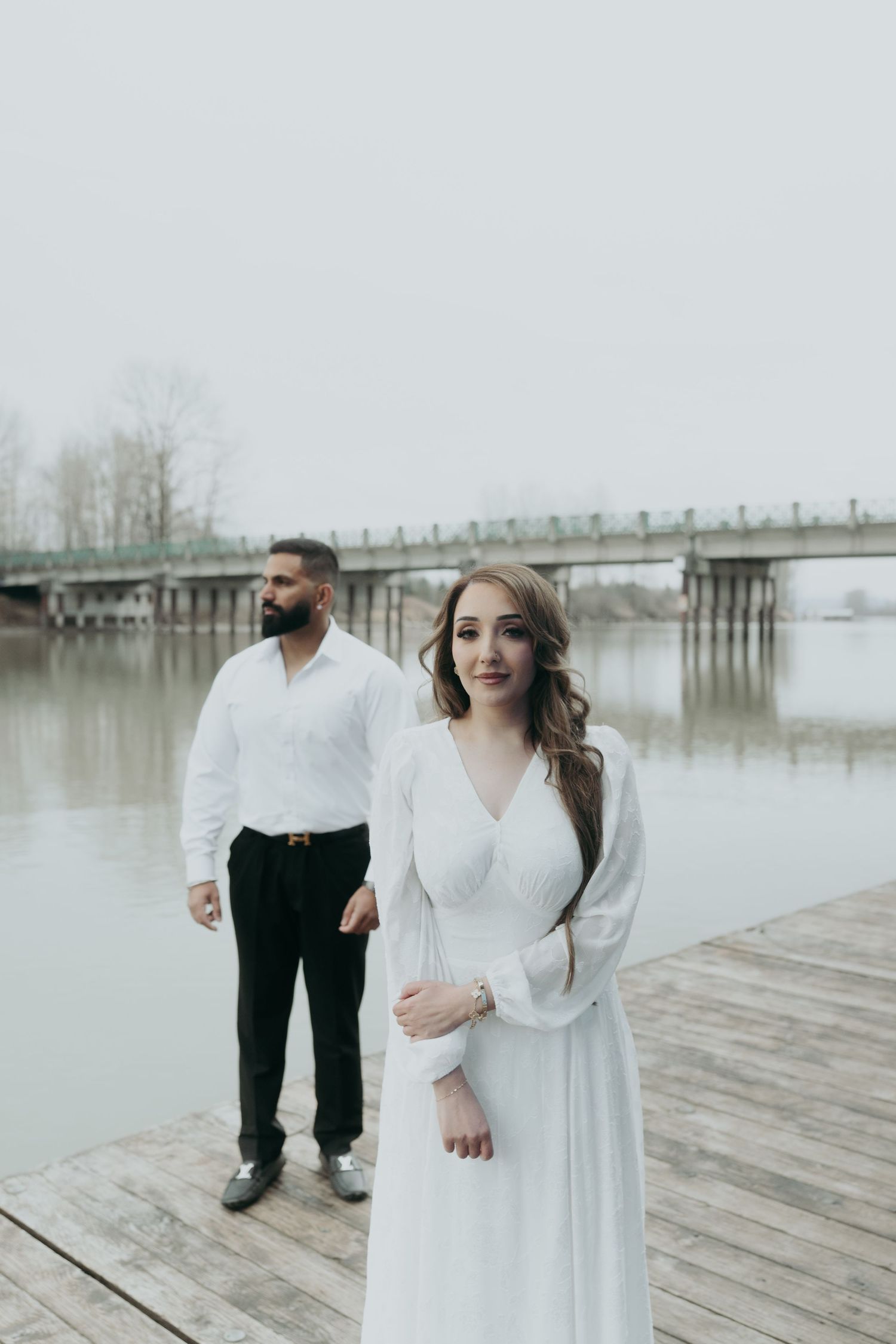 A couple poses on a misty wooden dock with a bridge in the background, wearing elegant white and black attire.