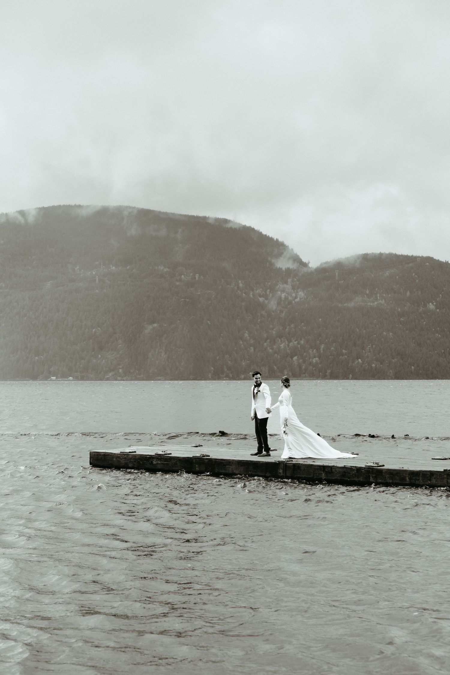 A couple stands on a wooden dock overlooking a misty lake surrounded by mountains in black and white photography.