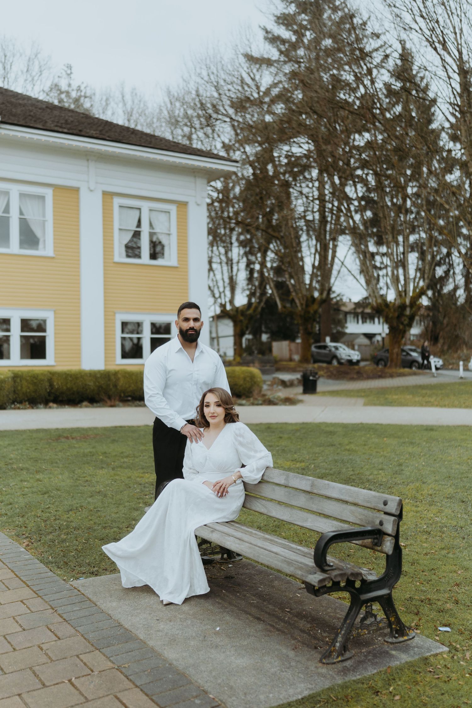 A romantic portrait of a couple by a park bench with a yellow building in the background.