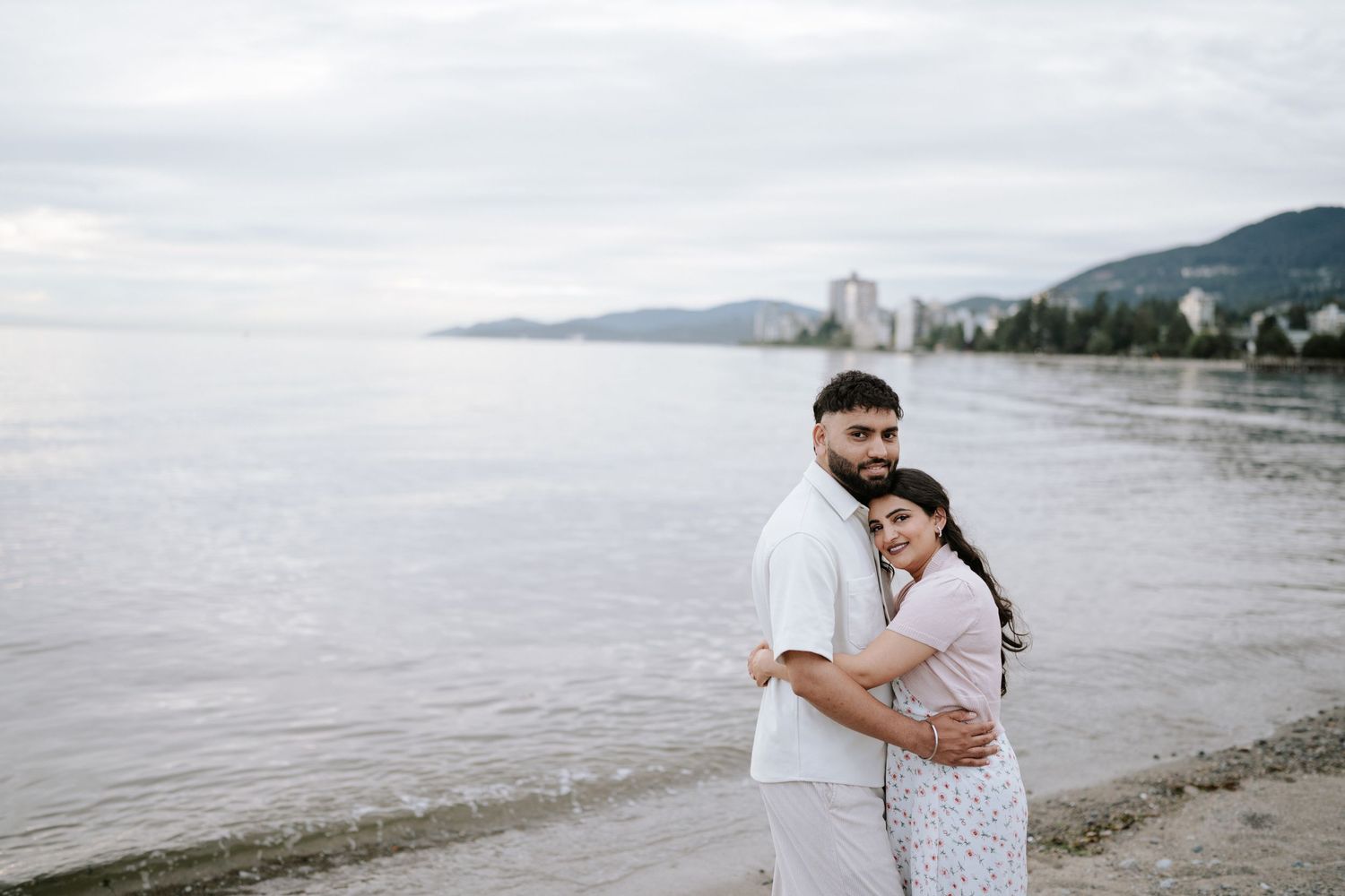 A couple in coordinated white outfits shares a tender moment on a scenic beach.