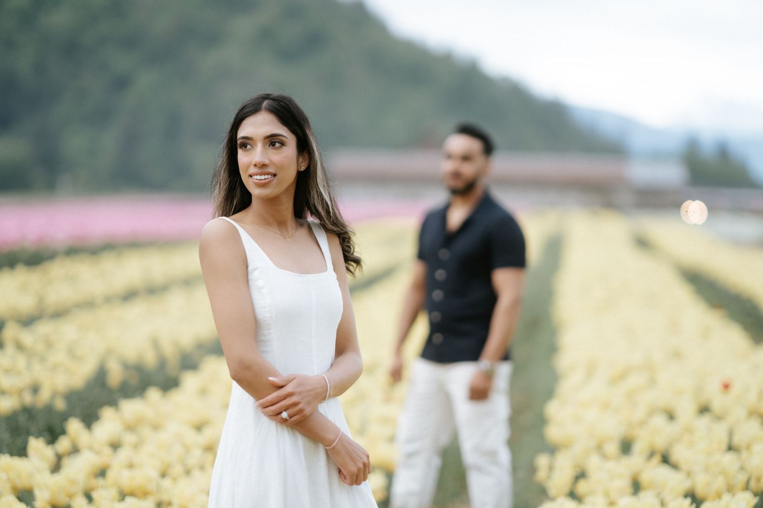 Two people stand in a scenic field of yellow flowers with mountains visible in the background.