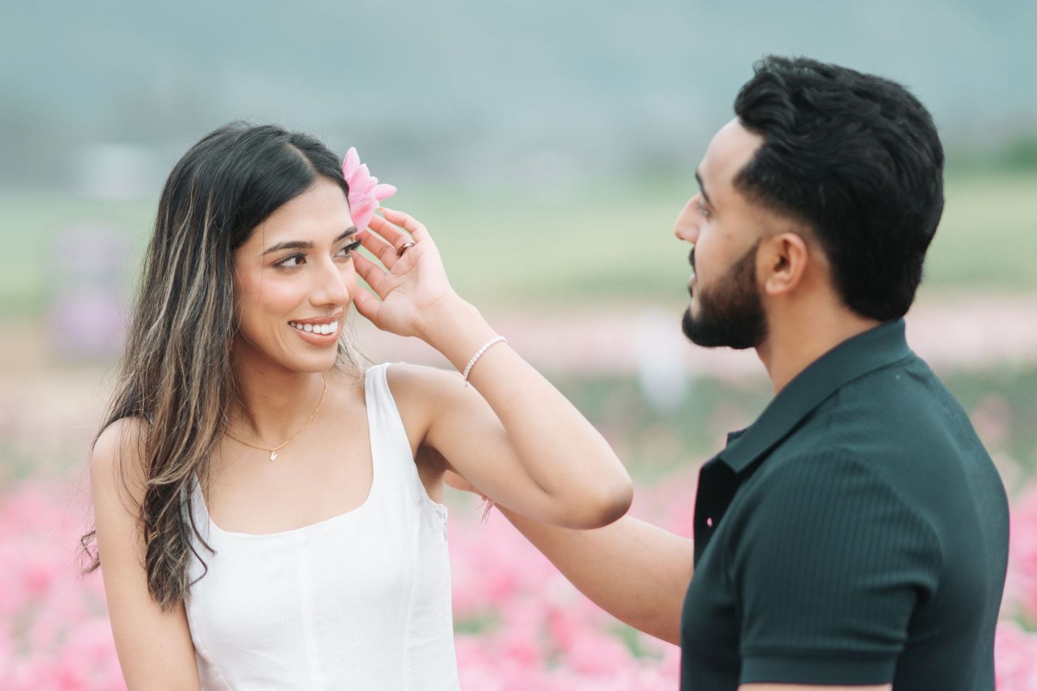 A couple shares romantic moments in a pink flower field while dressed in wedding attire during a photo session.