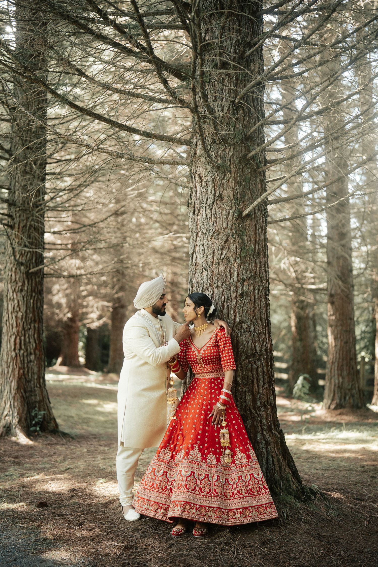 Wedding couple posing against tall pine trees in traditional Indian wedding outfits.