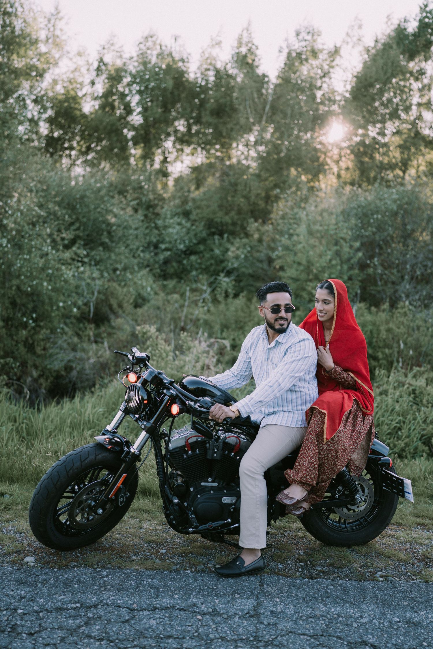 A couple sits on a custom black motorcycle on a gravel path surrounded by green foliage at dusk.