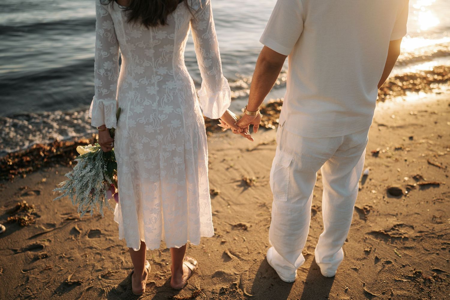Couple wearing white outfits walking together on a sandy beach at sunset with waves in the background.