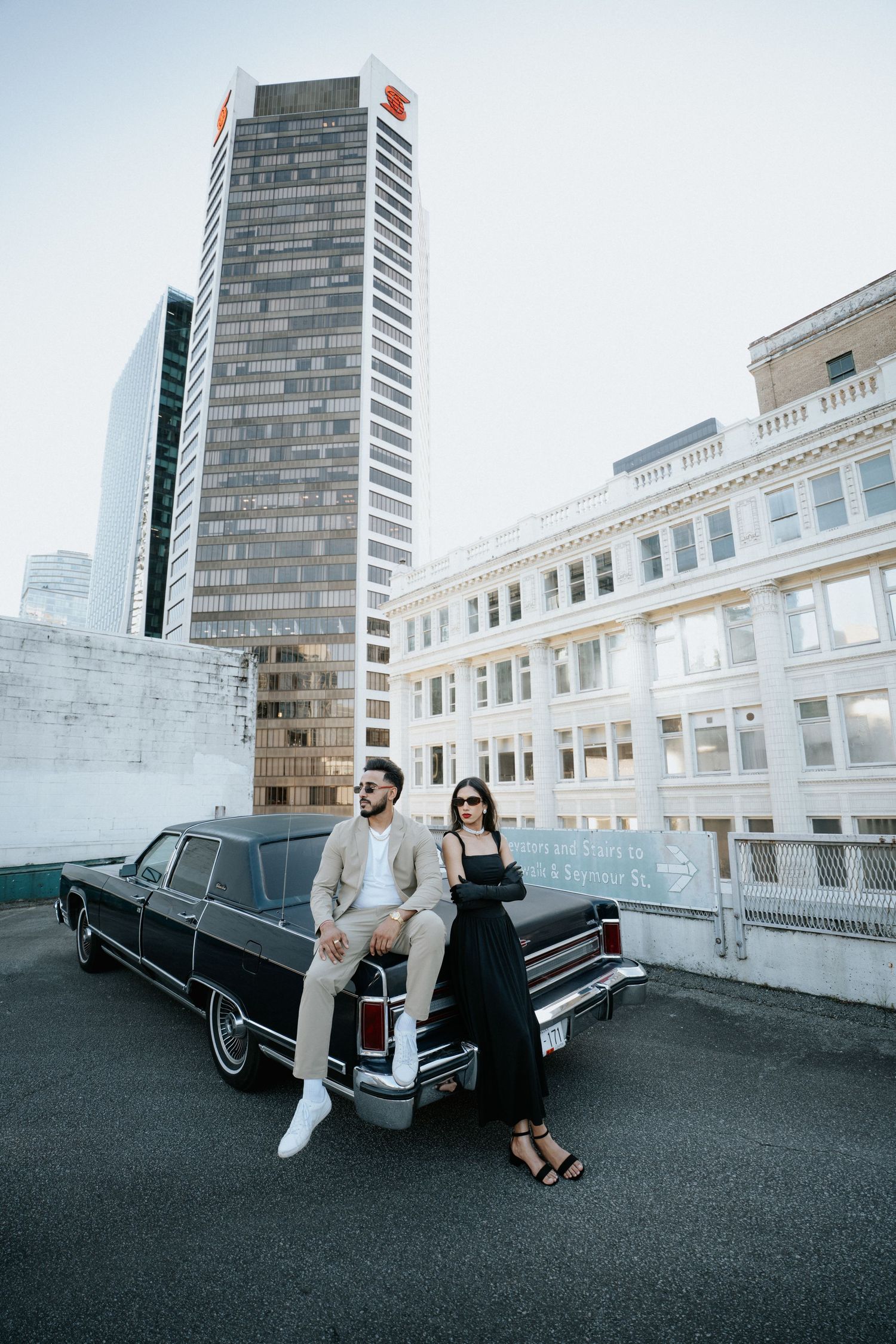 A couple poses next to a vintage black car on a rooftop with city buildings in the background.